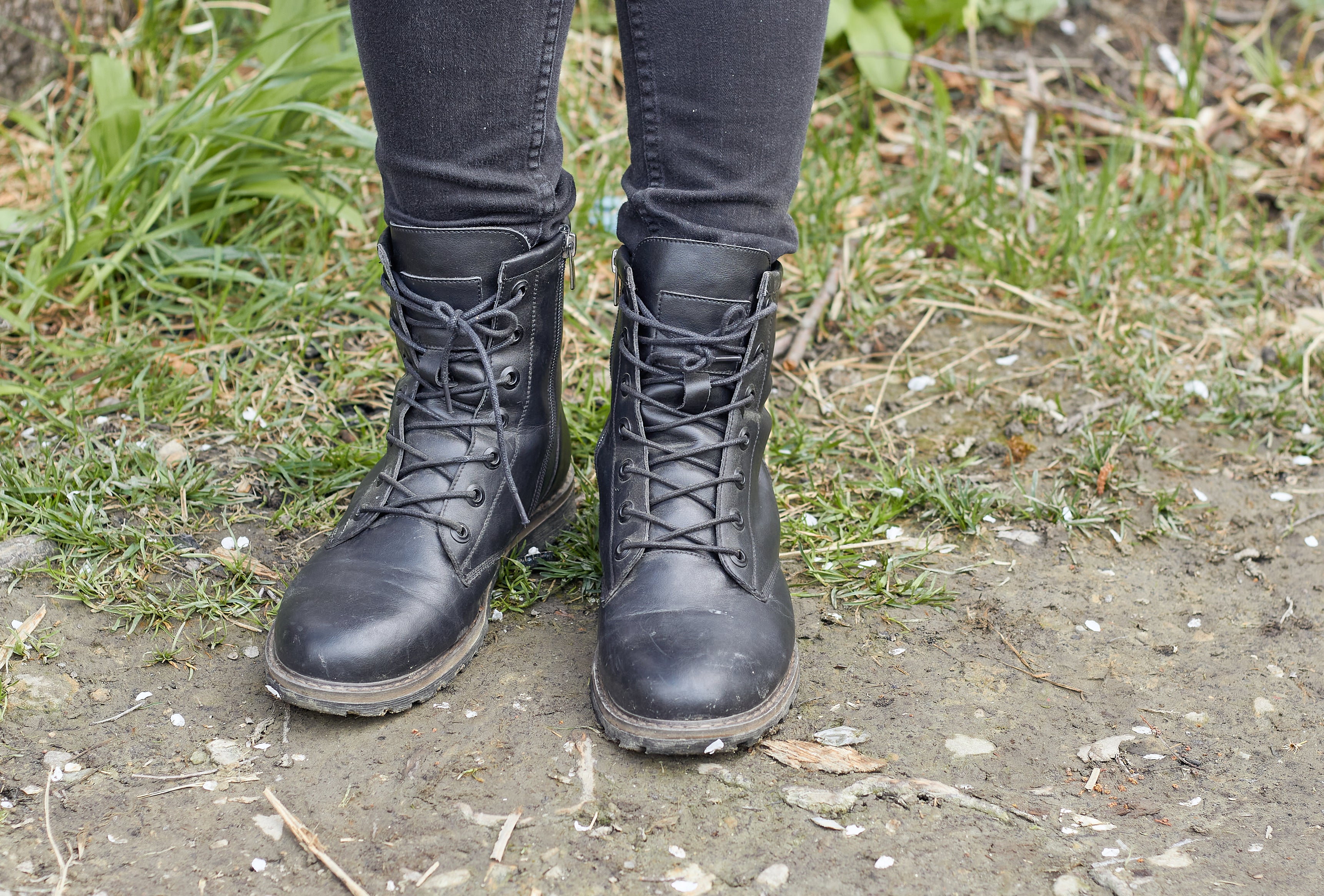 Person standing outdoors on uneven ground, shown from the knees down, wearing dark jeans and black lace-up boots