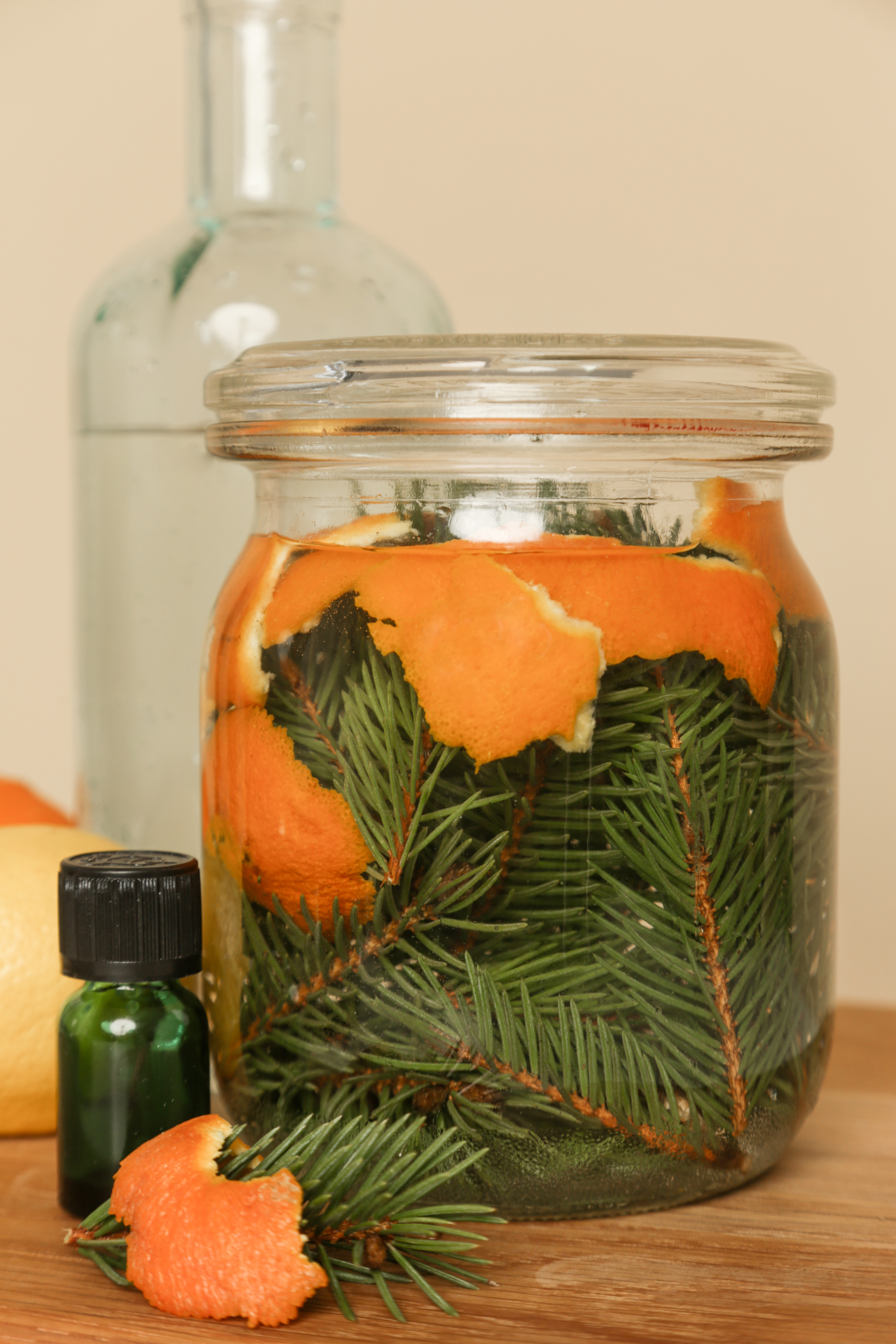 A glass jar filled with pine needles and orange peels next to a small green vial and an empty glass bottle in the background