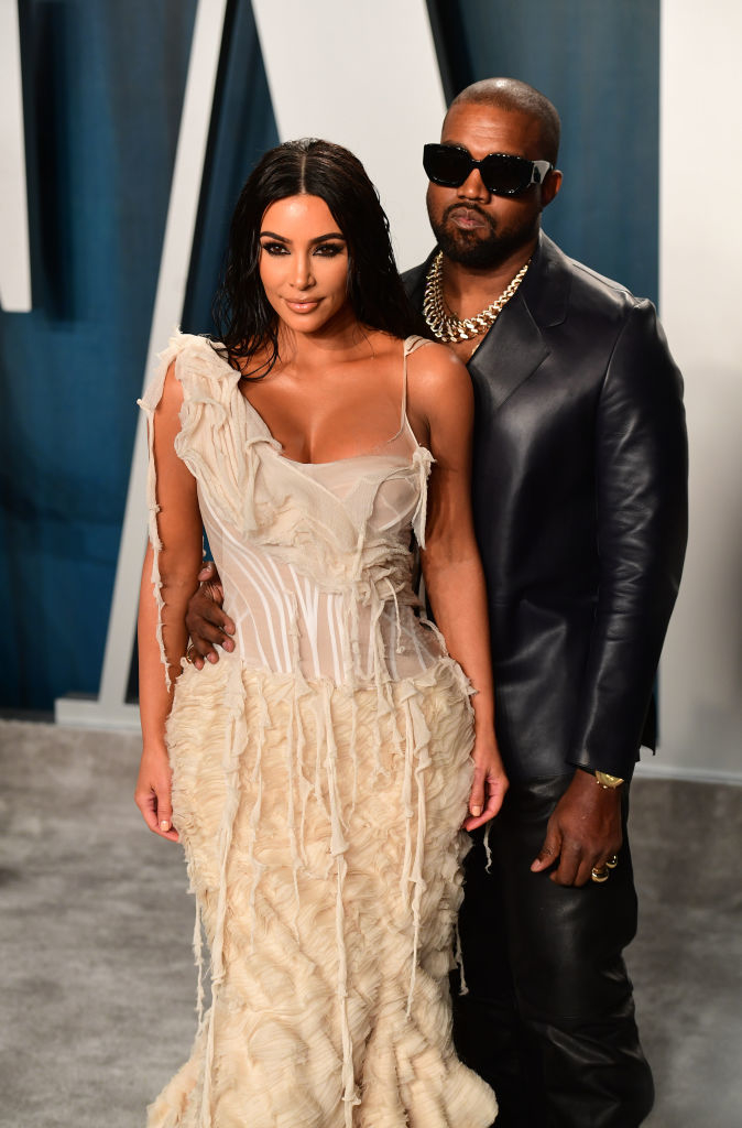 Kim Kardashian in a flowing textured gown stands next to Kanye West, who is wearing a dark blazer, gold chain, and sunglasses