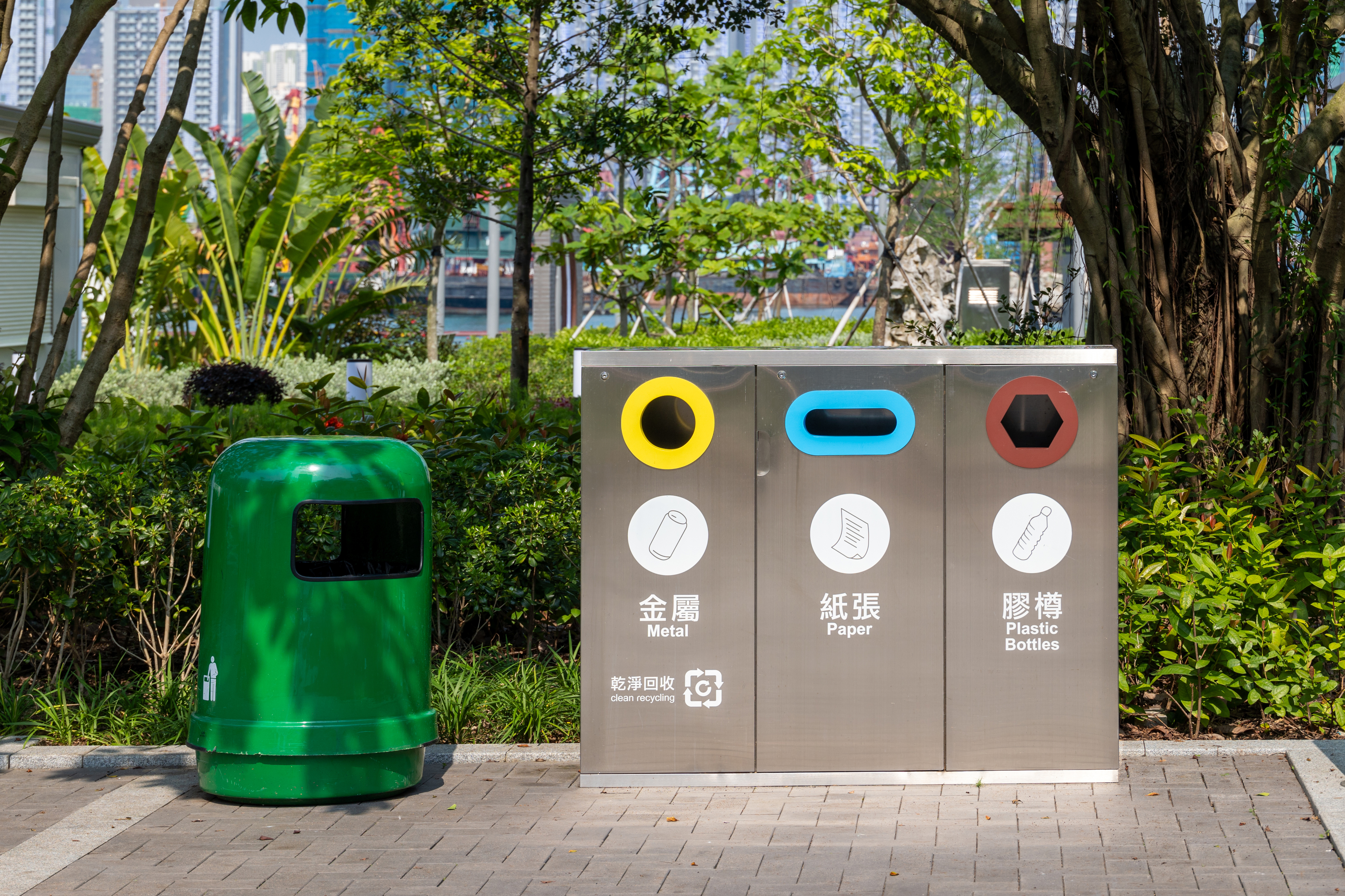 A public recycling station with bins for metal, paper, and plastic bottles next to a green garbage can in a park setting with trees and buildings in the background