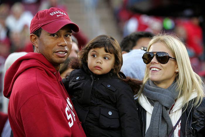 Tiger Woods, Elin Nordegren, and their child are bundled up in casual attire, outdoors at an event. Elin wears large sunglasses and a scarf