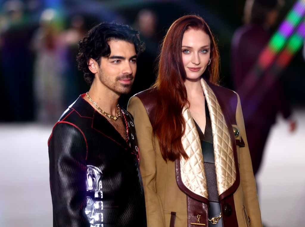 Joe Jonas and Sophie Turner stand together on a red carpet. Sophie is wearing a stylish coat with quilted details. Joe is dressed in a fashionable jacket