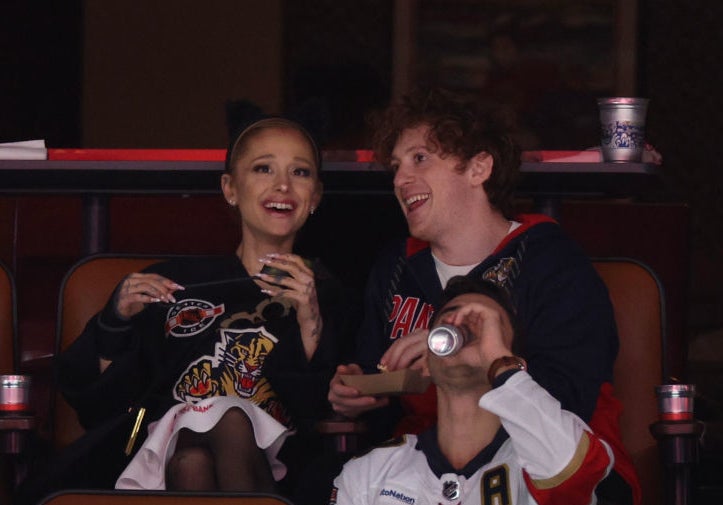 Ariana Grande and Dalton Gomez sit with friends in a stadium box, enjoying a hockey game. Ariana gestures while smiling, and Dalton wears a Florida Panthers jersey