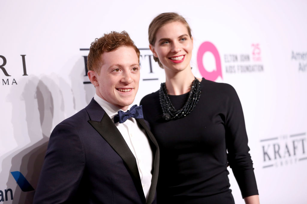 A man in a dark suit with a blue bow tie and a smiling woman in a black dress with a beaded necklace stand on a red carpet at a formal event