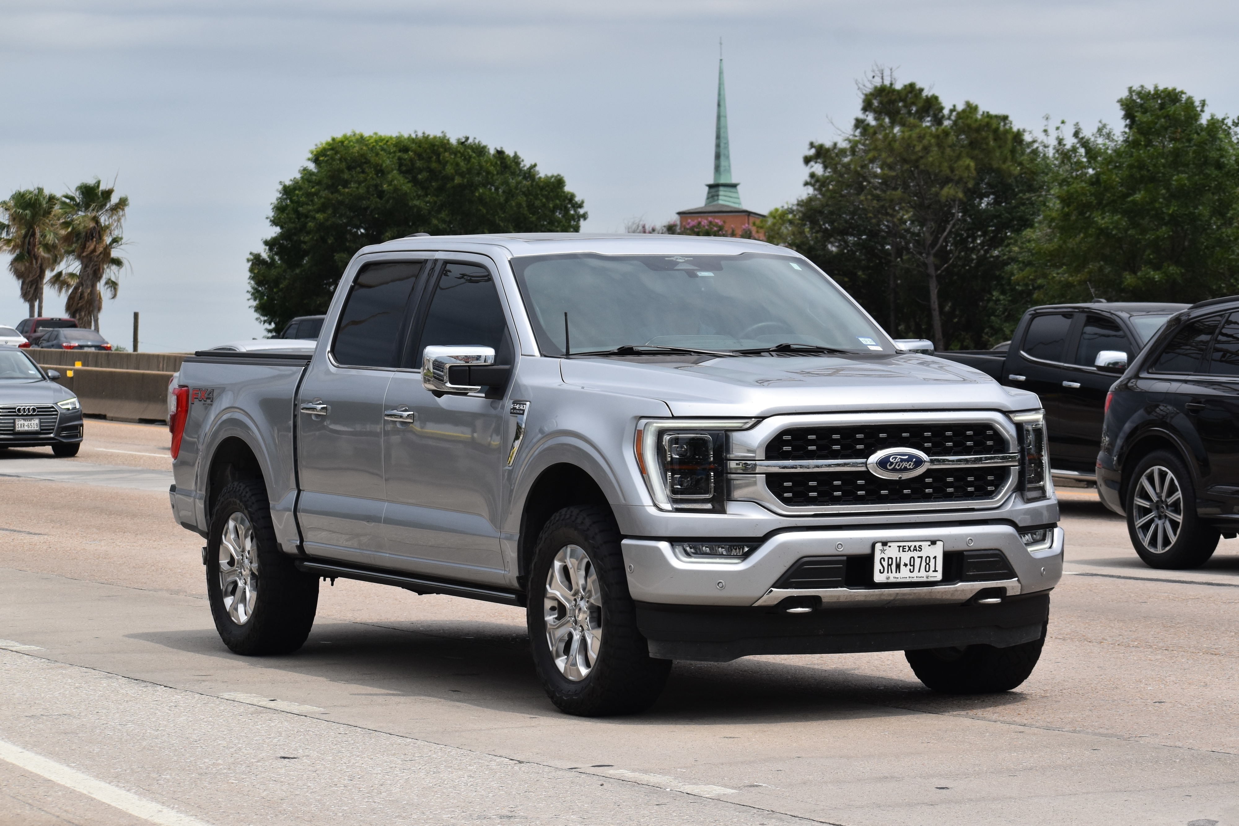 A silver Ford F-150 pickup truck driving on a road with other vehicles in the background. Trees and a building with a spire are visible. Texas license plate is visible