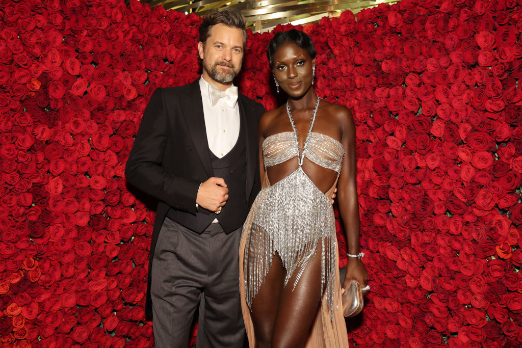 A man in a tuxedo and a woman in a glamorous, silver cut-out gown with a sheer train stand together in front of a wall of red roses