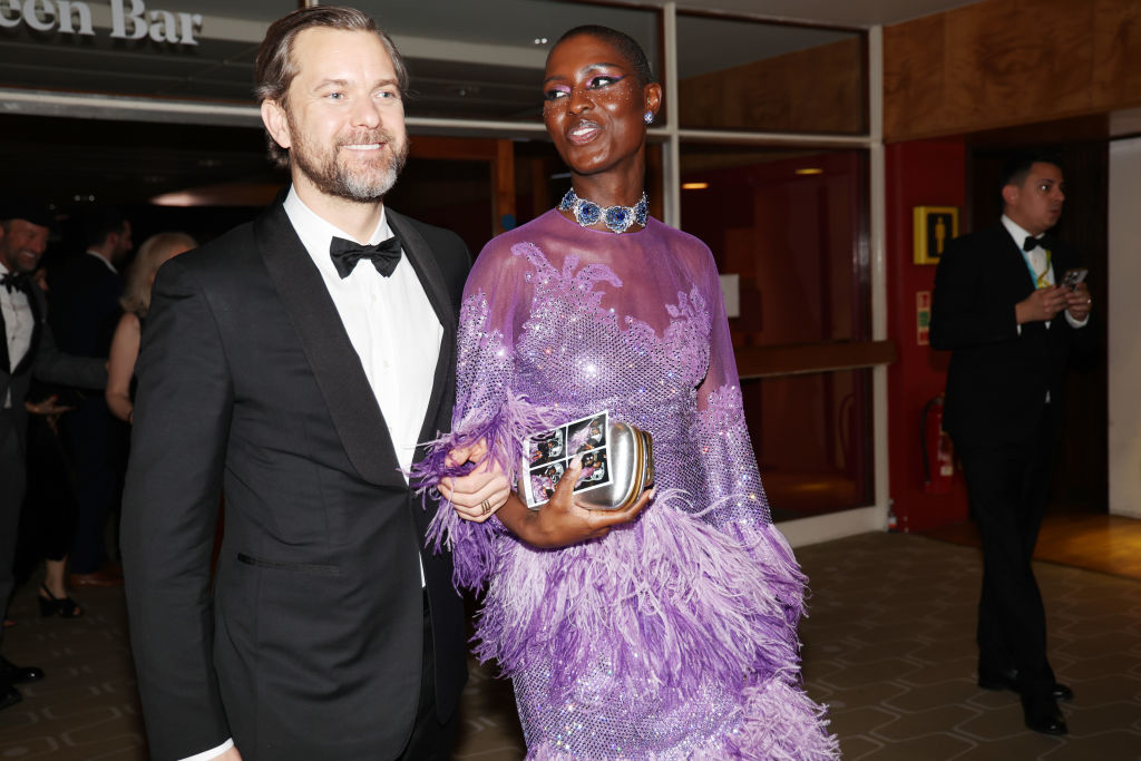 A man in a classic black tuxedo and a woman in a chic, feathery purple gown hold hands at an event