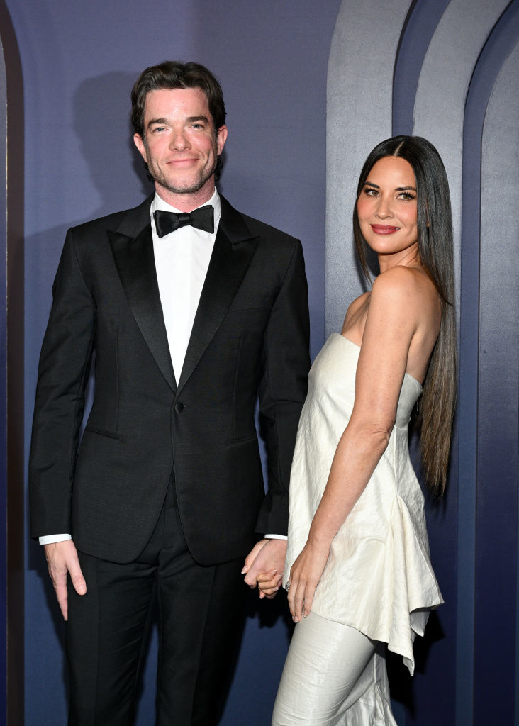 John Mulaney in a tuxedo and Olivia Munn in an off-shoulder top with pants, holding hands and posing together