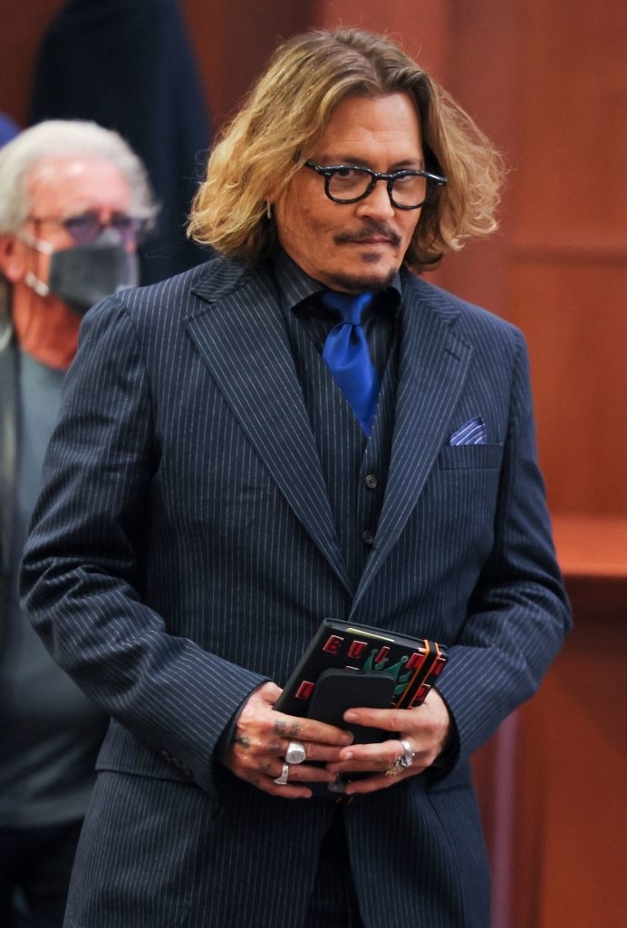Johnny Depp in a pinstripe suit, blue tie, and glasses, holding a notebook. An individual wearing a mask is visible in the background