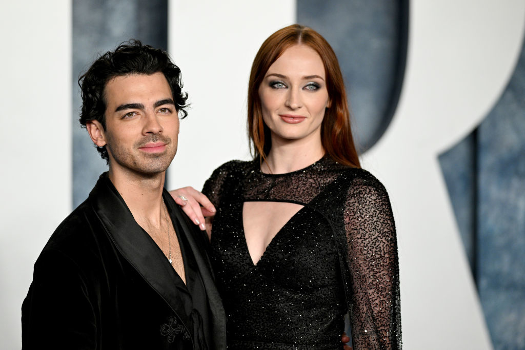 Joe Jonas and Sophie Turner pose together. Joe wears a dark blazer over a low-cut shirt, and Sophie wears a black sequined dress with sheer sleeves