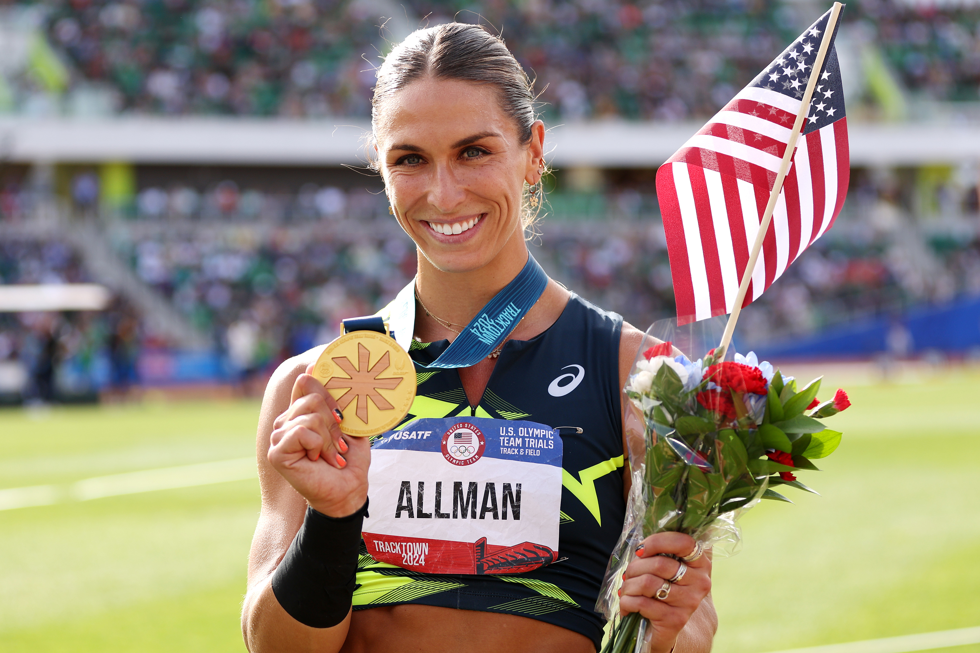 Athlete Valarie Allman holds a gold medal and an American flag, wearing an athletic top with her name displayed, and smiles at the camera