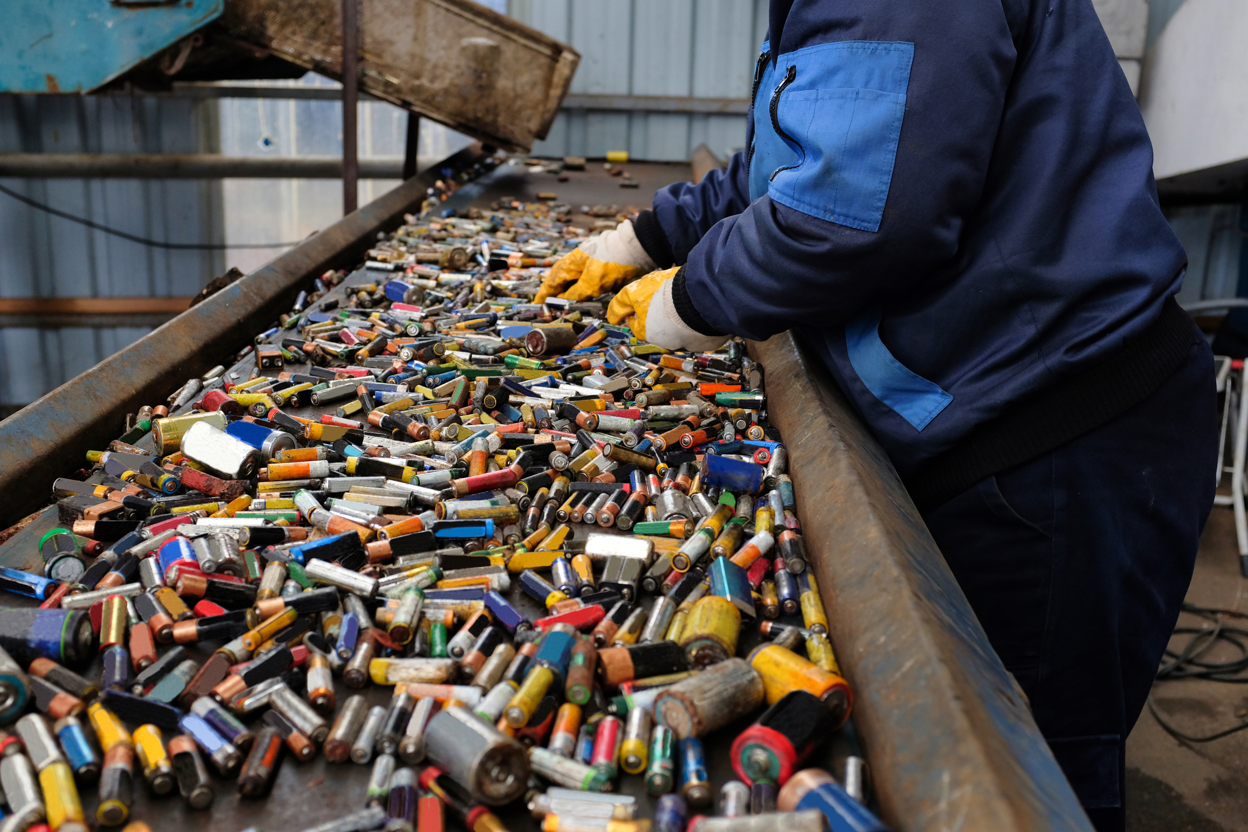 Person in a blue jacket and gloves sorts various types of batteries on a conveyor belt in a recycling facility