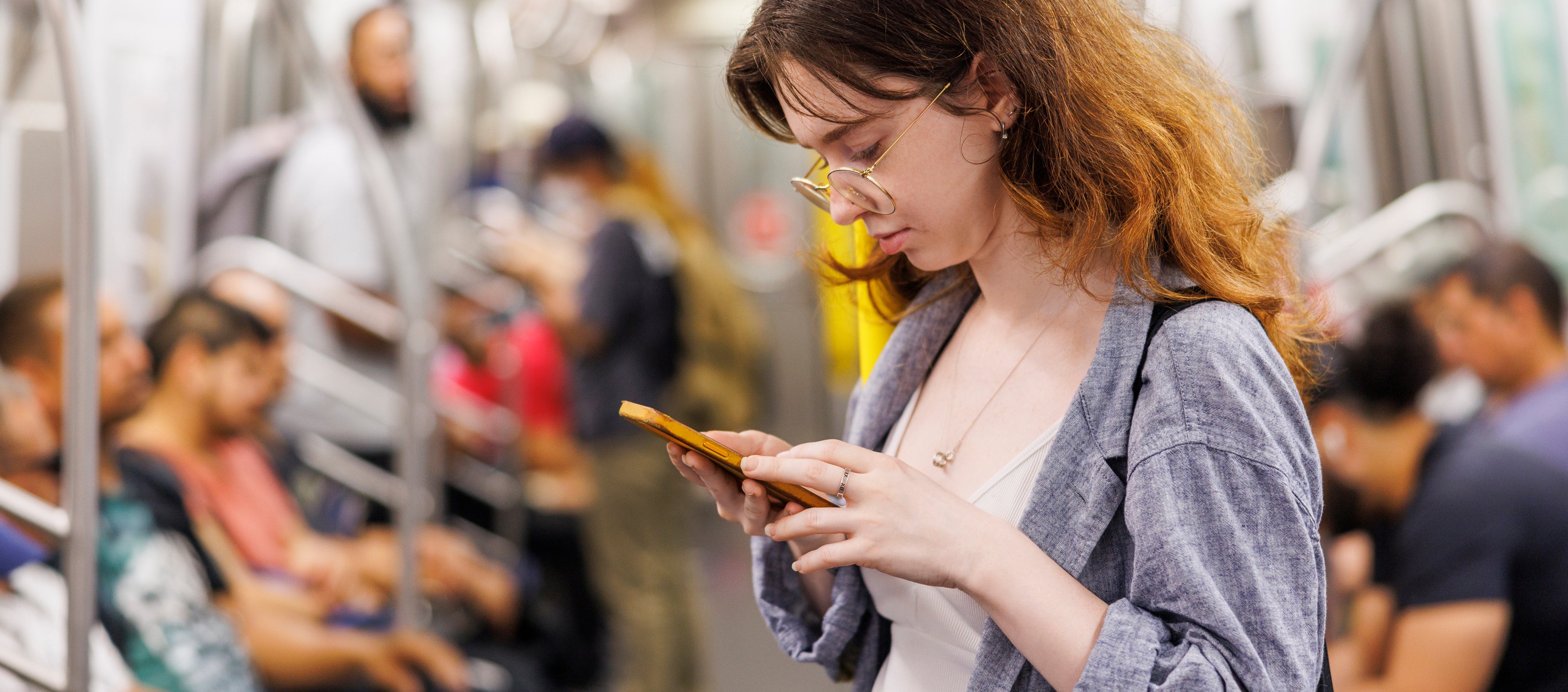 A woman stands in a subway car, focused on her phone. Other passengers are seated around her, all engaged with their own activities
