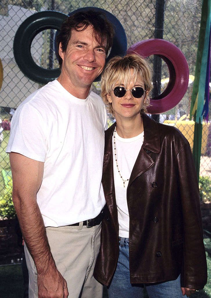 Dennis Quaid in a white T-shirt and beige pants with Meg Ryan in a leather jacket, white T-shirt, jeans, and sunglasses, smiling at an outdoor event