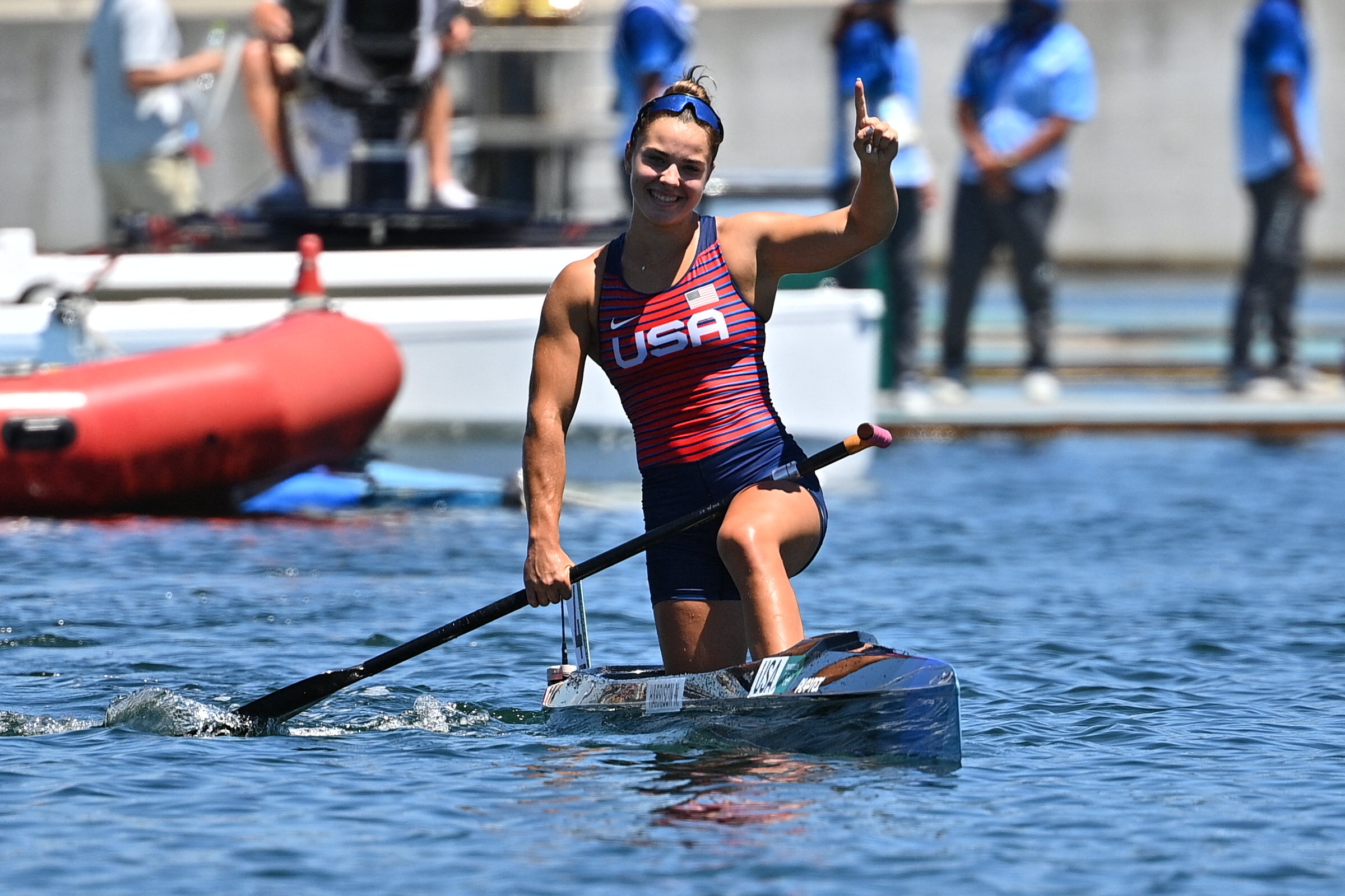 Olympic canoe athlete paddles, smiling and raising number one finger while wearing USA uniform. Other crew members in blue shirts blurred in the background