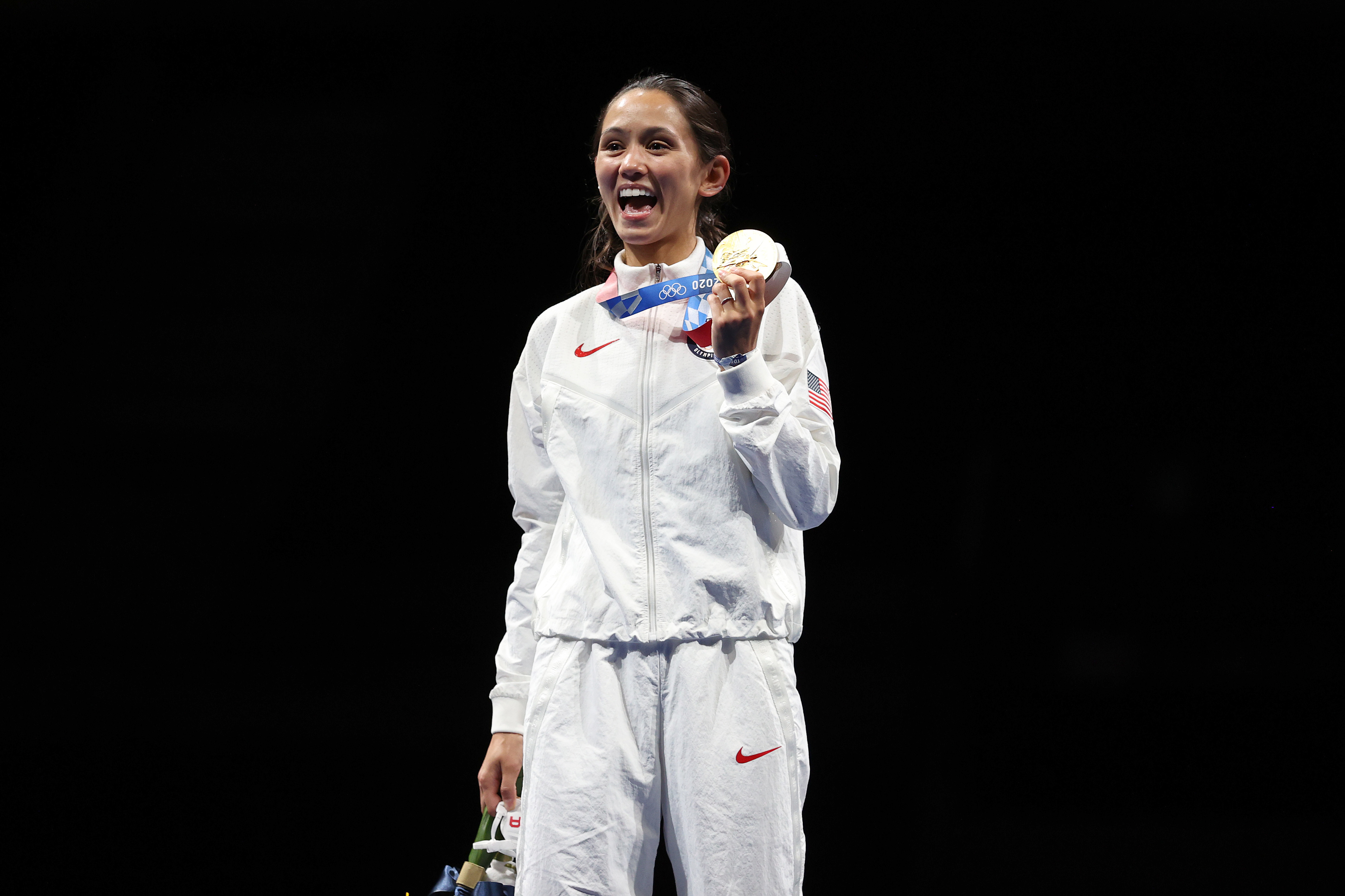 An athlete in a white tracksuit holds a medal and a bouquet, smiling widely on a podium