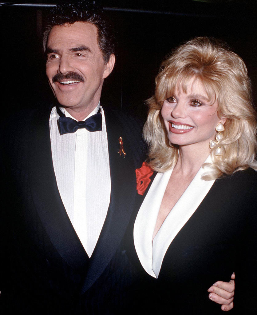 Burt Reynolds and Loni Anderson smiling, posing together. Reynolds is in a tuxedo with a bow tie, Anderson is wearing a plunging neckline dress with pearl earrings