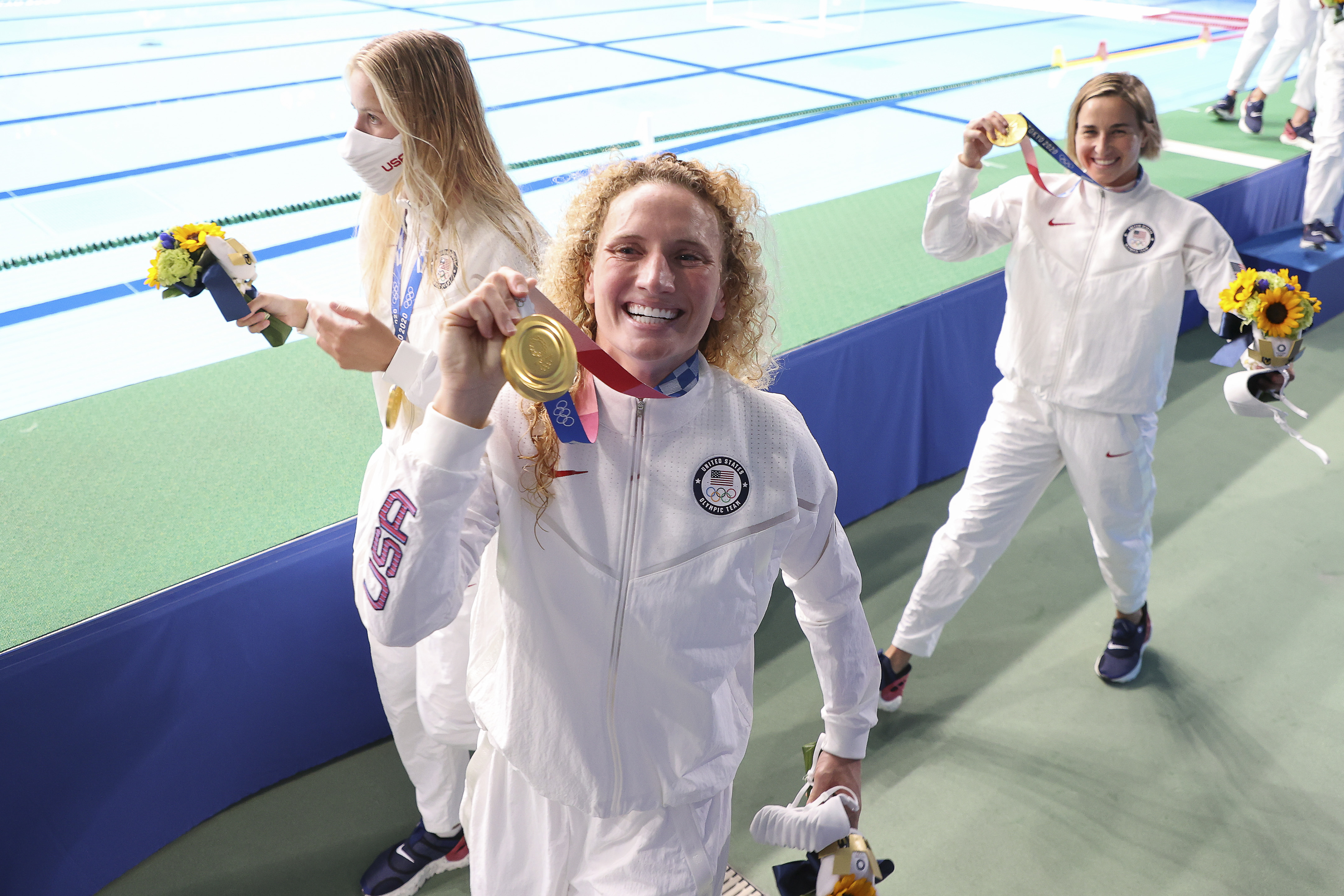 Swimmers with gold medals at the event, including one holding a bouquet, pose victoriously in athletic suits. Swimming lanes are visible in the background
