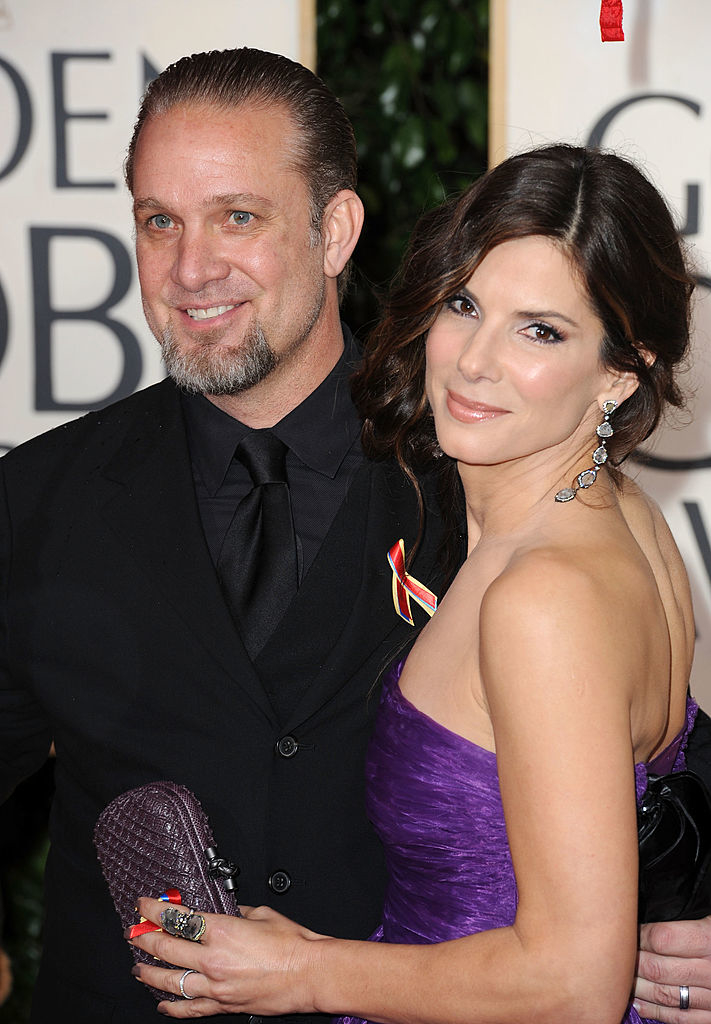 Jesse James and Sandra Bullock pose together on a red carpet, with Sandra in a strapless gown and holding a clutch, and Jesse in a black suit with a ribbon pin