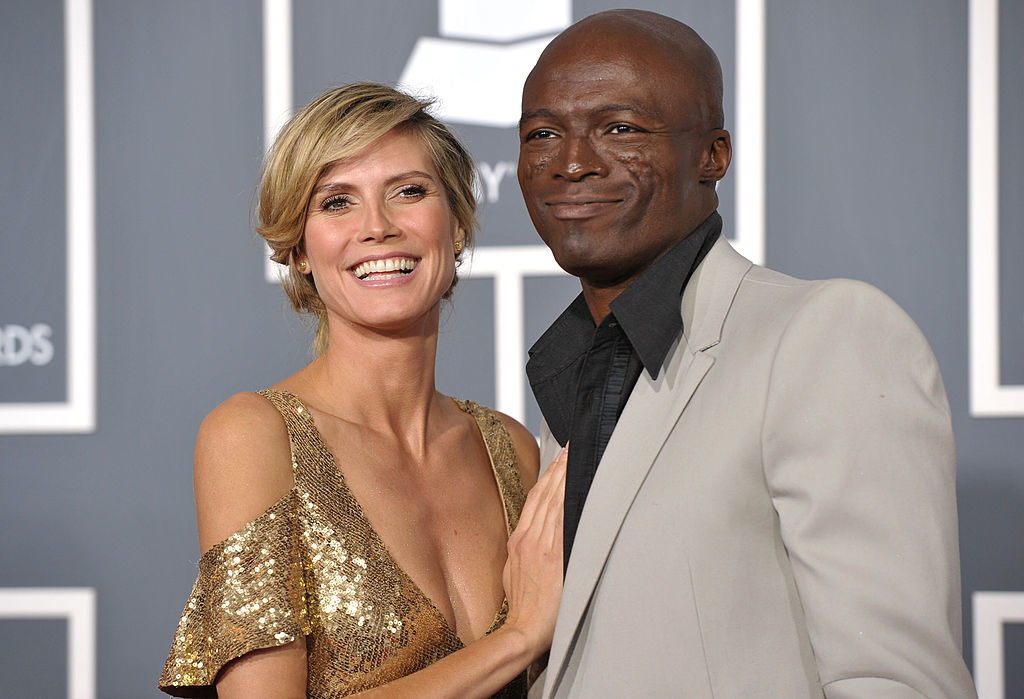 Heidi Klum in a sparkling, sequined dress stands smiling next to Seal, who is wearing a light-colored suit and dark shirt, at an event