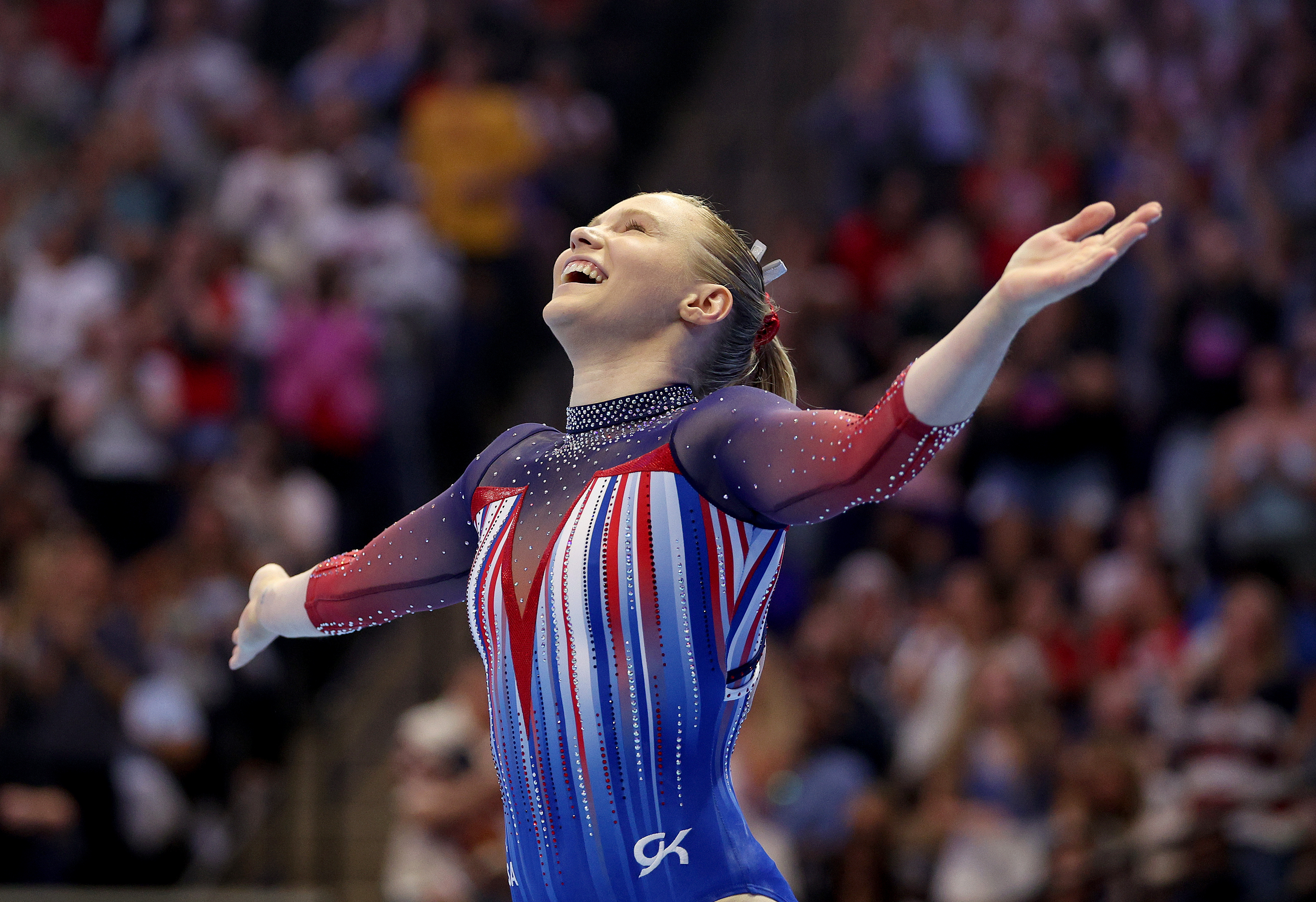 Gymnast smiling with arms outstretched, wearing a blue leotard with red, white, and striped patterns, likely during a performance at a sports event