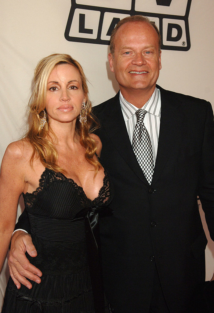 A man and woman, Kelsey Grammer and Camille Grammer, posing together on a red carpet. Camille is wearing a black dress, and Kelsey is in a suit and tie