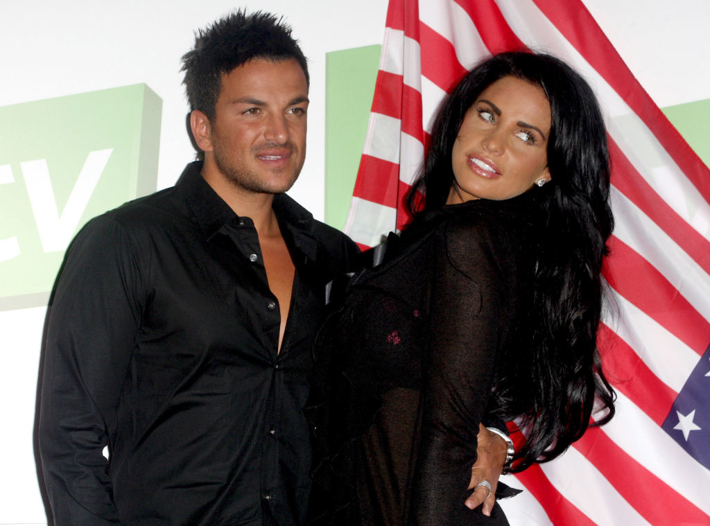 Peter Andre and Katie Price pose in front of an American flag backdrop. Peter wears a black shirt, Katie wears a stylish black outfit