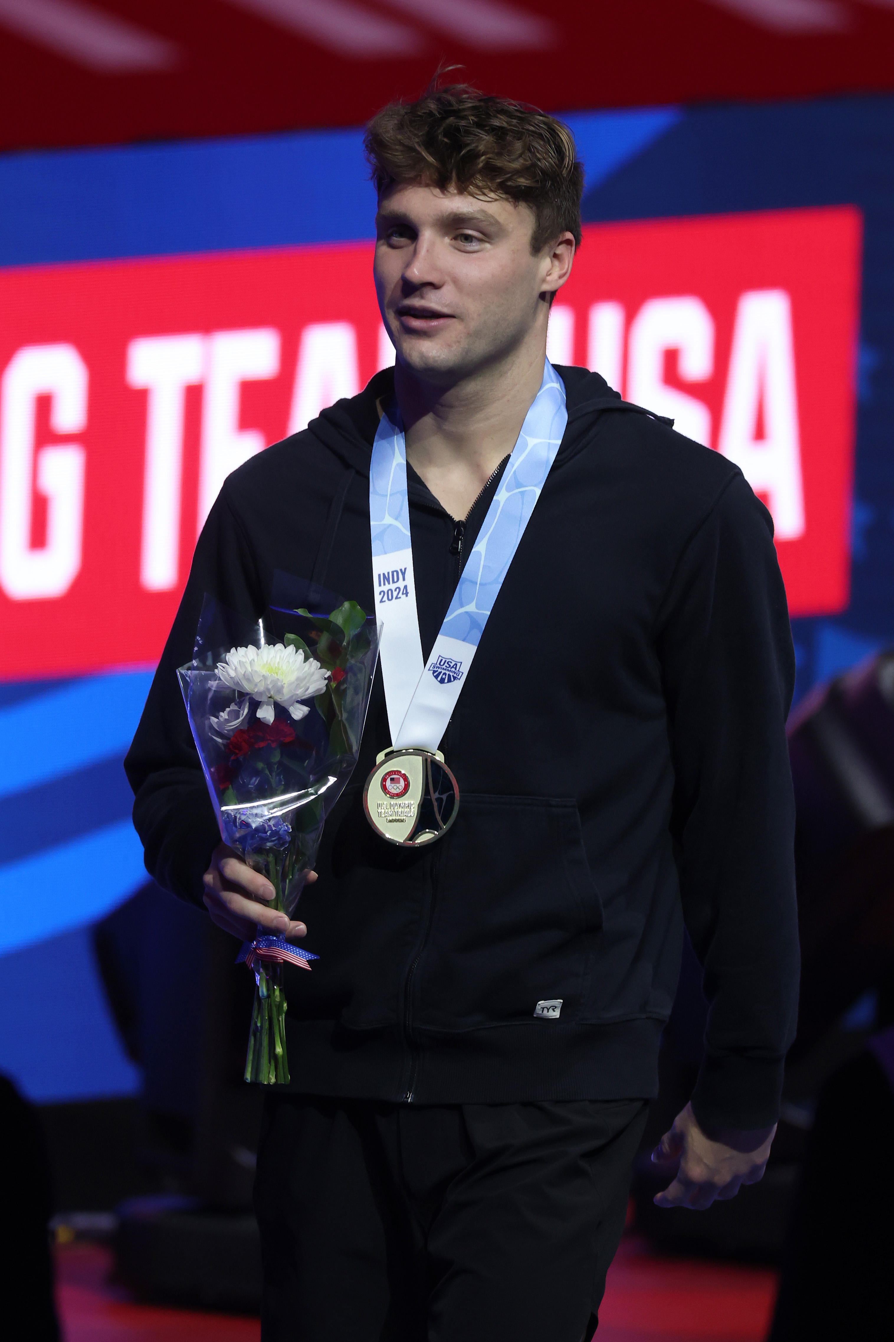 Bobby Finke in black casual outfit holds a bouquet and medal in front of a &quot;Team USA&quot; backdrop at a sports event