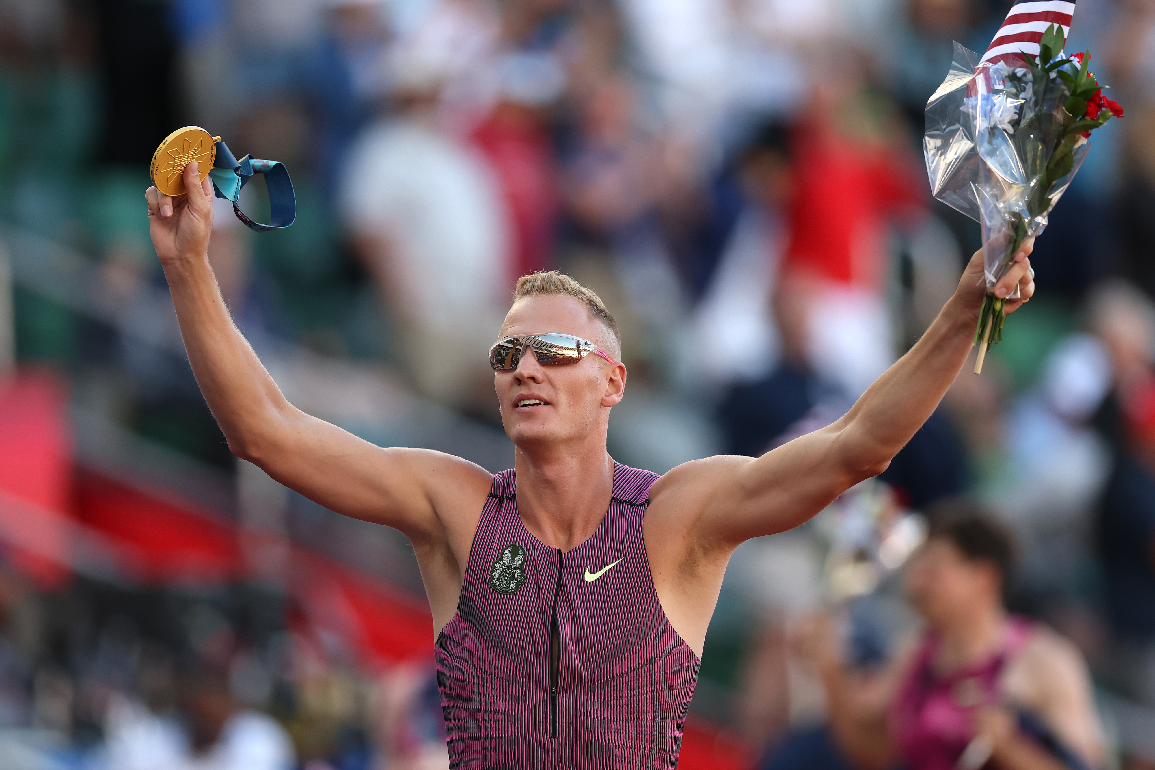 Athlete holds a gold medal and a bouquet, raising his arms in celebration at a sports event