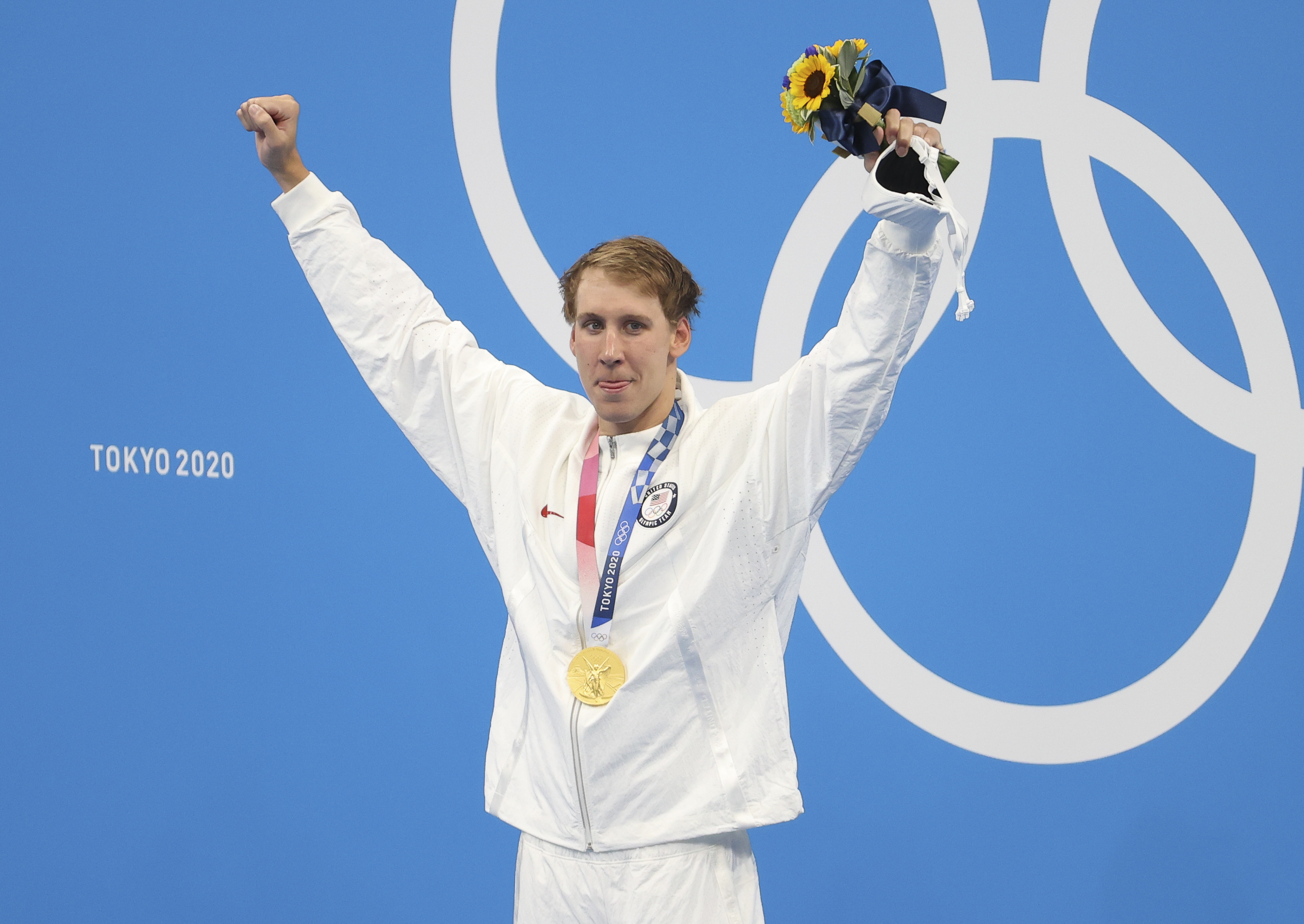 Olympic swimmer stands on podium with gold medal and flowers, arms raised in victory, against Tokyo 2020 backdrop
