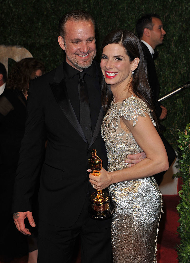 Sandra Bullock in a shimmering dress holds an Oscar, standing arm-in-arm with a man in a suit at an event. Both are smiling warmly