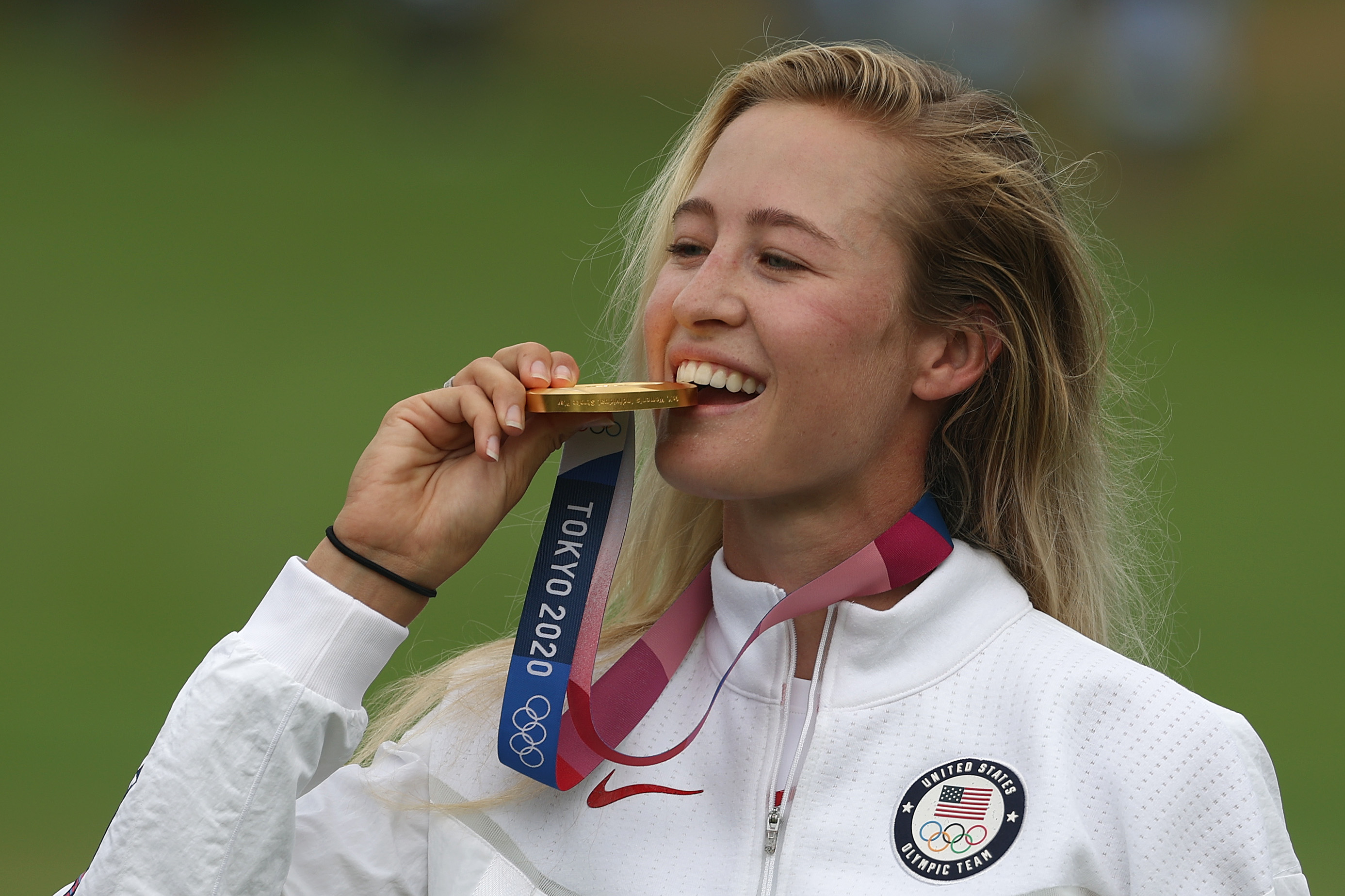 Athlete Nelly Korda bites her gold medal while wearing a white USA Olympic Team jacket at the Tokyo 2020 Olympics