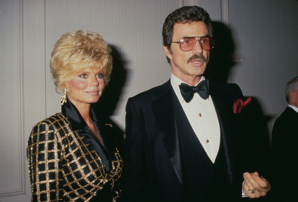 Loni Anderson and Burt Reynolds at an event, Loni in a patterned jacket and Burt in a tuxedo with a bow tie and tinted glasses