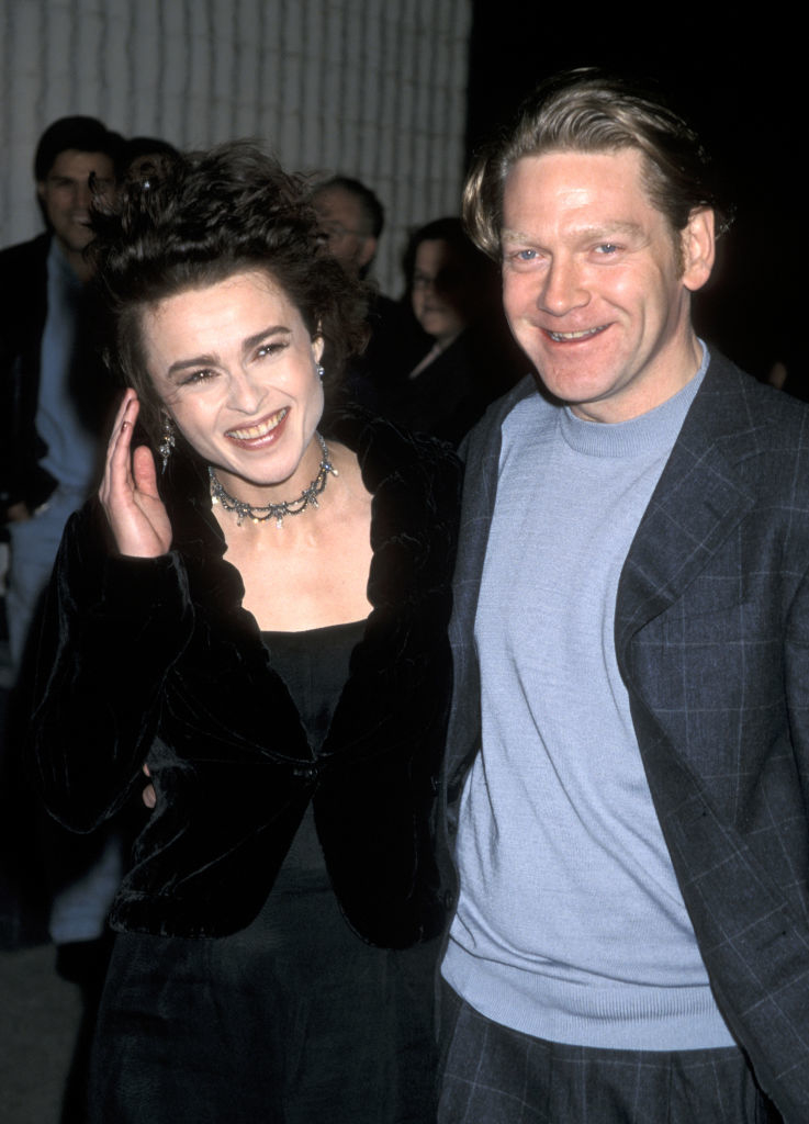 Helena Bonham Carter, in a black velvet outfit with a ruffled neckline and a choker necklace, poses with Kenneth Branagh, who is wearing a grey suit
