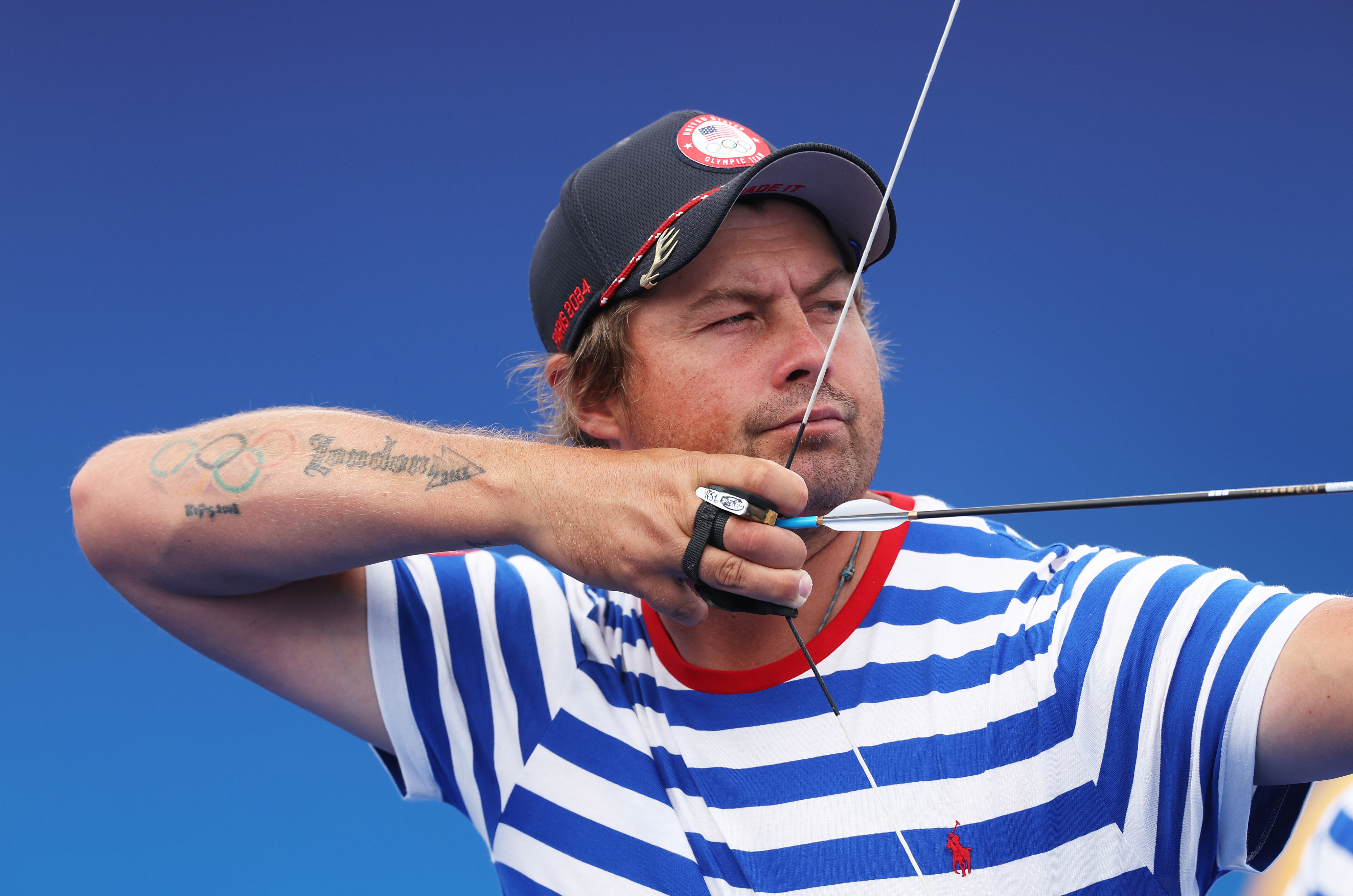 Archery athlete in a striped shirt and cap draws his bow, aiming intensely. Tattoo and rings visible on his arm and hand