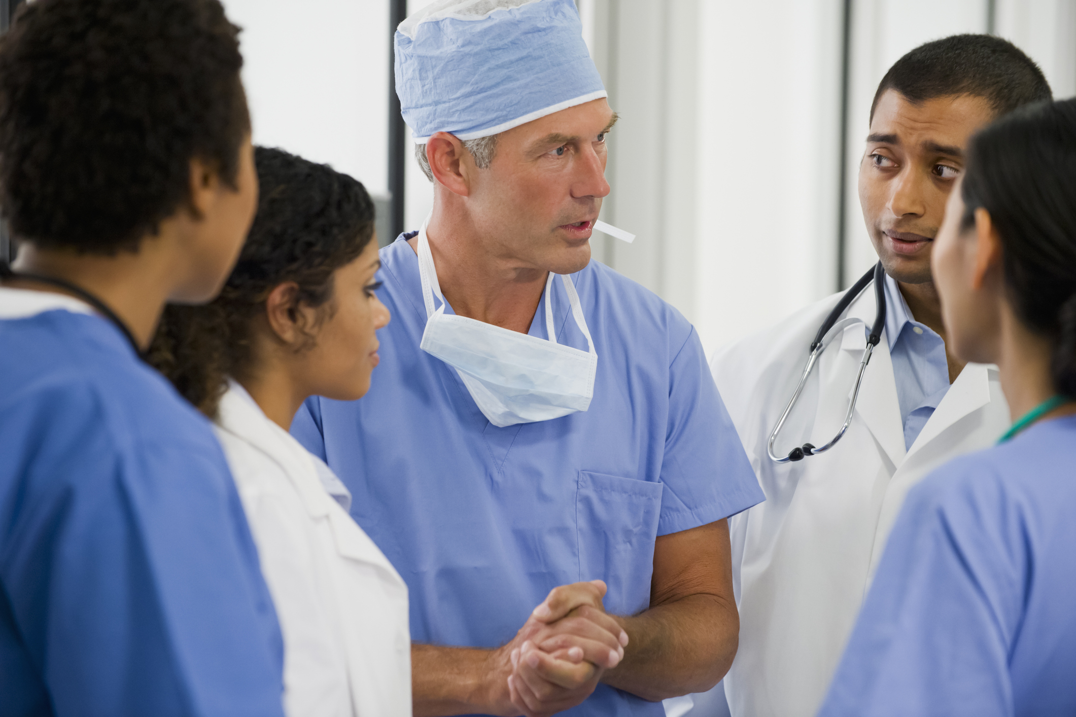 A group of medical professionals in scrubs and lab coats, including a surgeon with a cap and mask, engaged in a serious discussion