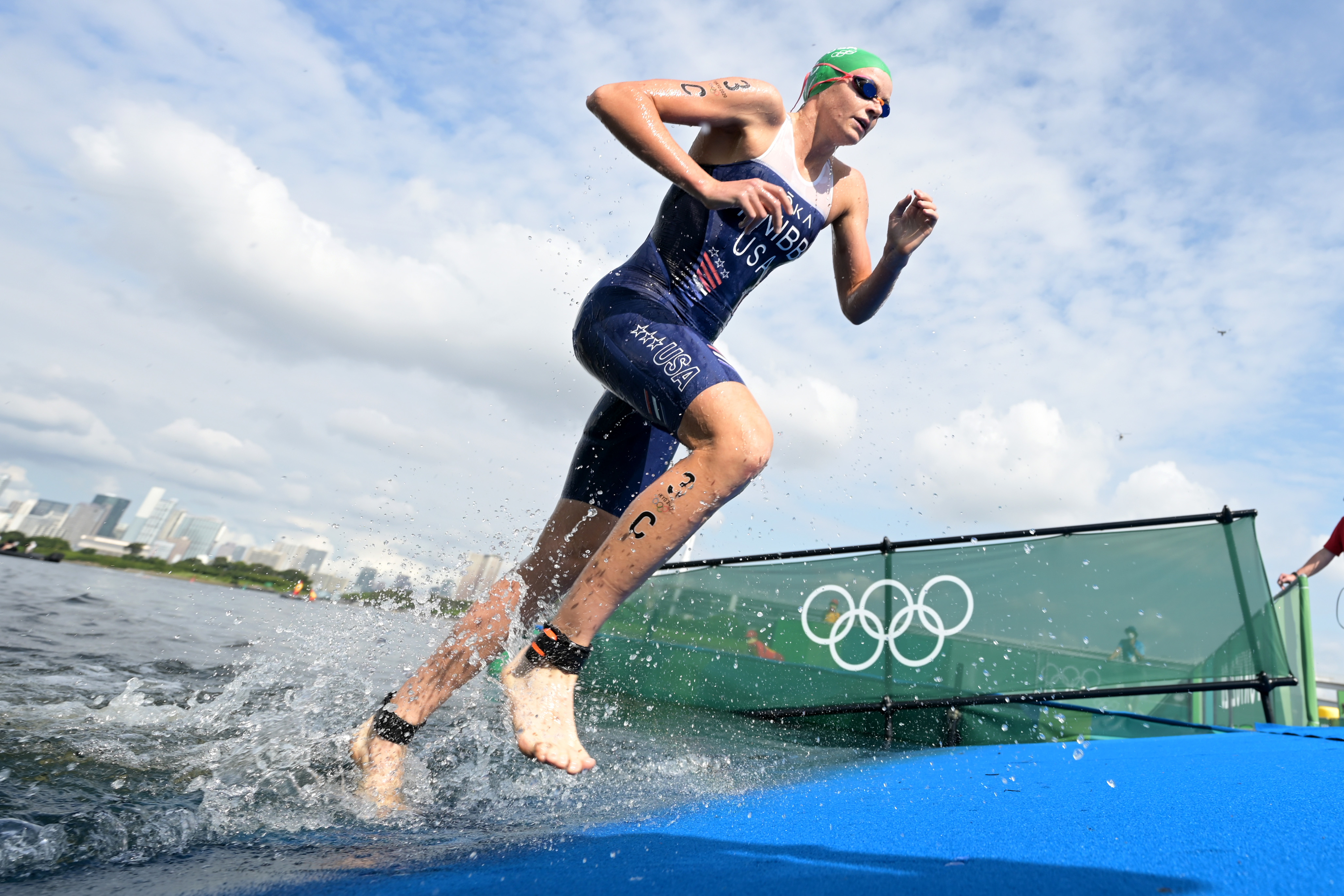 Taylor Knibb emerges from the water during an Olympic triathlon event, with splashing water and Olympic rings visible