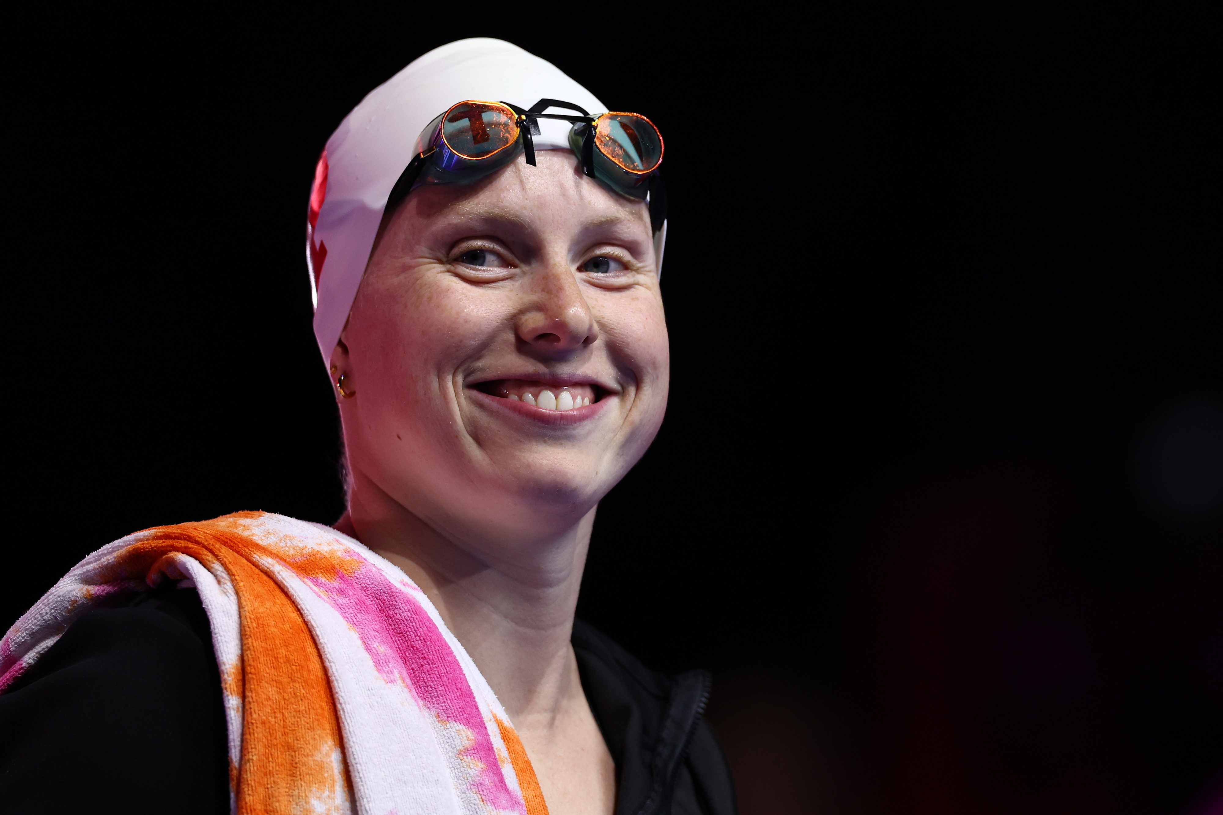 An unidentified smiling swimmer with a swim cap, goggles on forehead, and a towel draped over the shoulder. The image is categorized as Sports