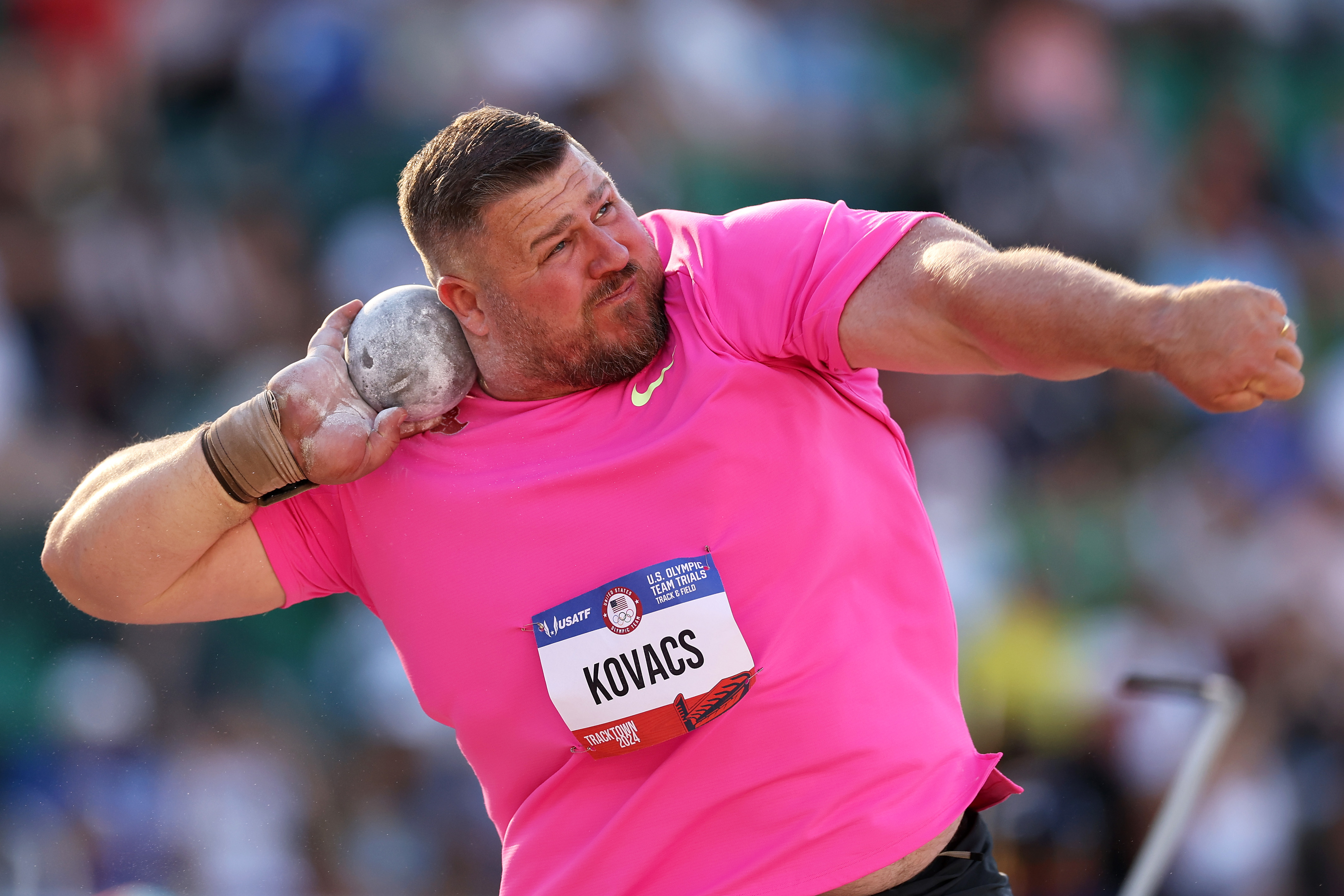 Joe Kovacs angles his body and prepares to throw a shot put during a sports event. He wears a bright T-shirt and has a determined expression