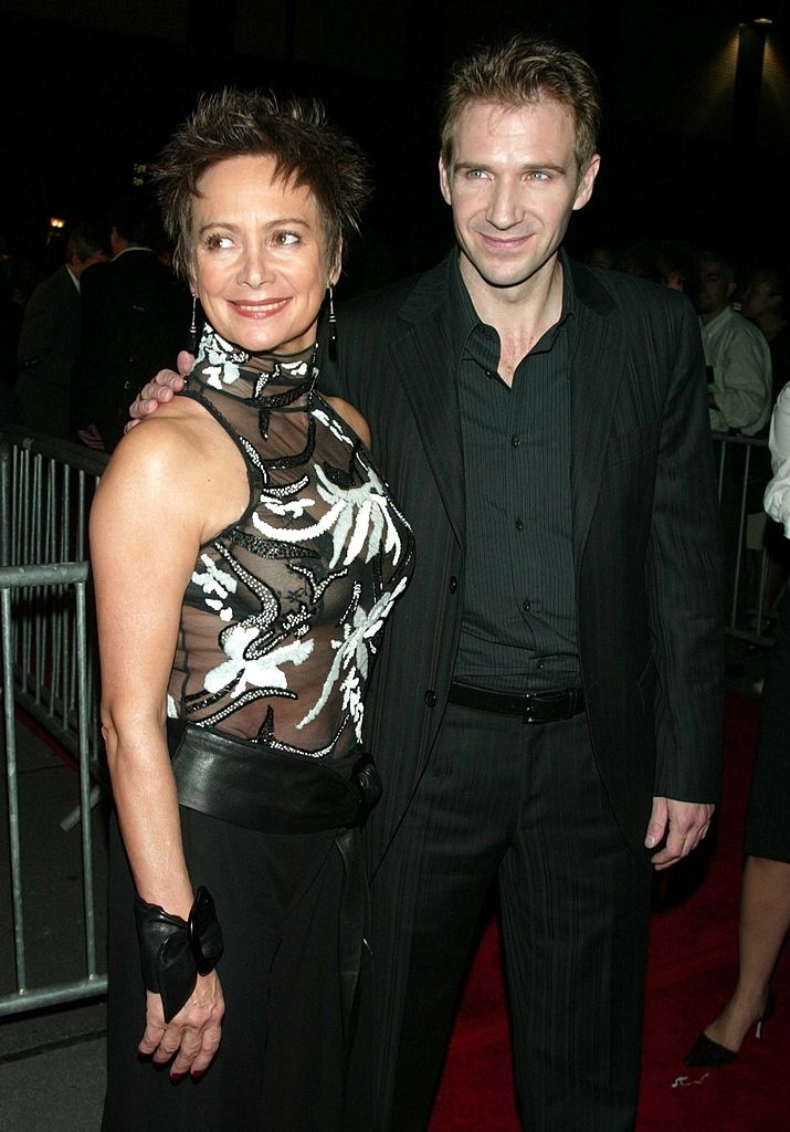 Two people, Ralph Fiennes in a black suit and Lindsay Duncan in an elegant sleeveless dress with floral detailing, stand together on a red carpet