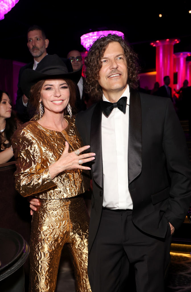Shania Twain in a gold, patterned suit and black hat, poses with a man in a black tuxedo at a formal event