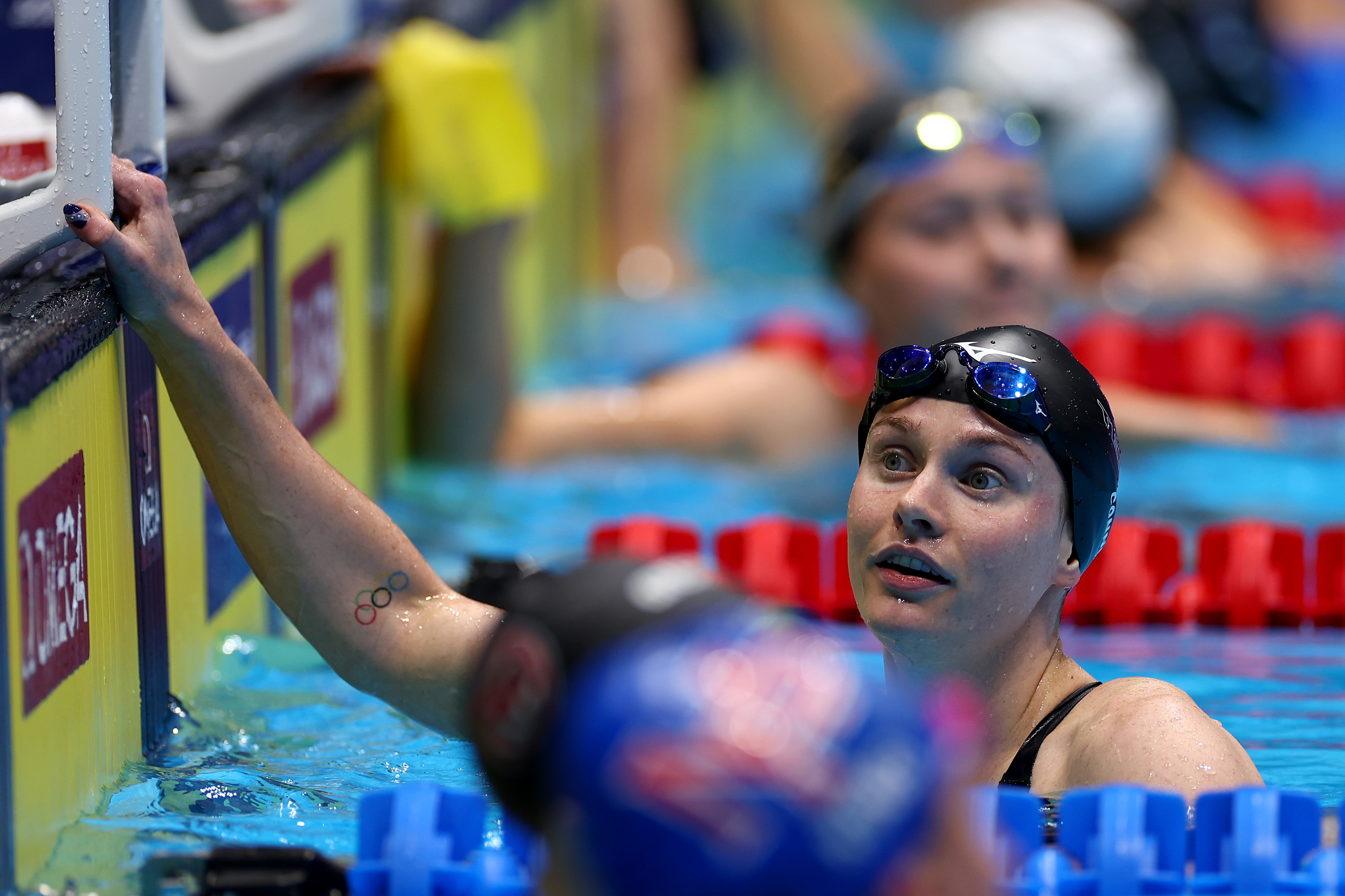 Swimmer in black cap with goggles, resting at pool edge, Olympic rings tattoo visible on arm, other swimmers in background