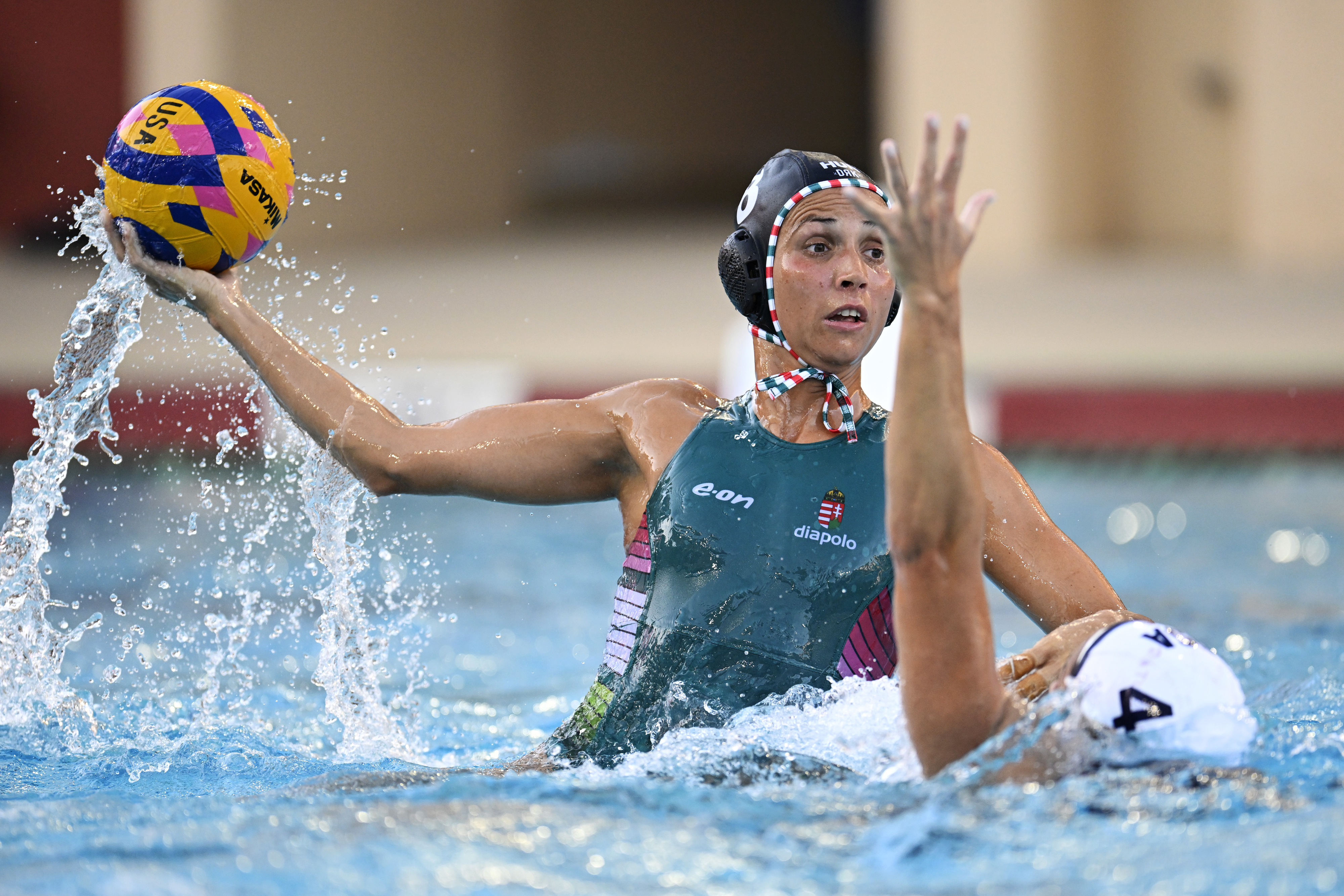 Two female water polo players, Rachel Fattal with the ball about to throw. Both are wearing swim caps and athletic swimsuits, in a dynamic pool setting