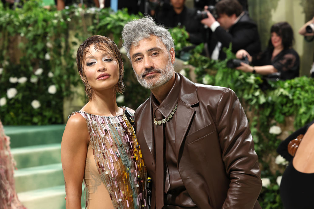 Rita Ora in a beaded, sleeveless gown and Taika Waititi in a brown leather suit pose together at a formal event