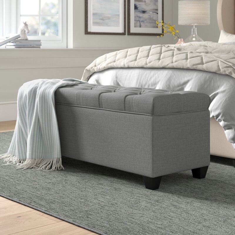 Bedroom scene with a gray padded storage bench at the foot of the bed