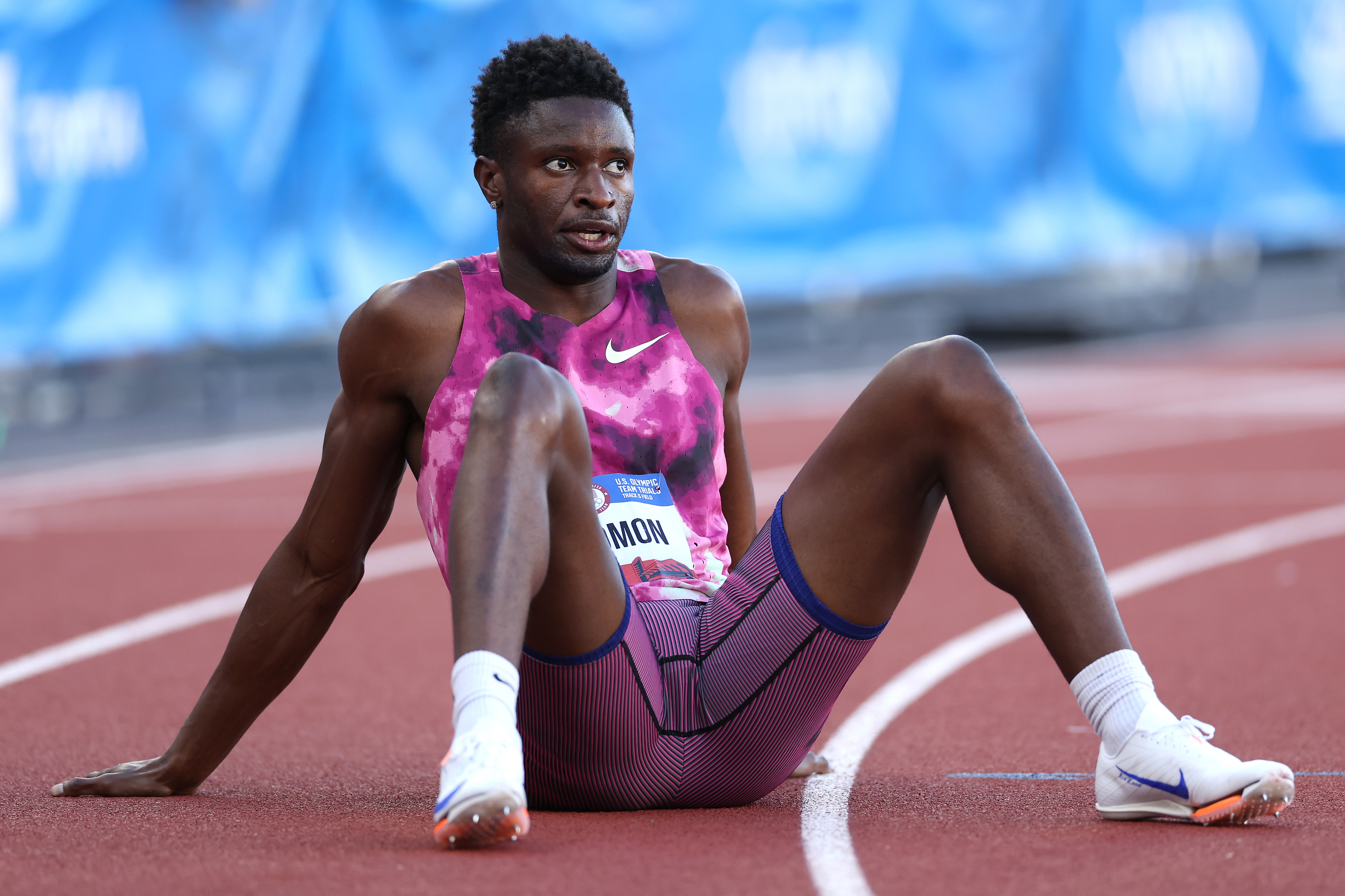 Athlete Rai Benjamin sits on the track after a race, wearing a sleeveless sports top and shorts, with a competition bib showing his name