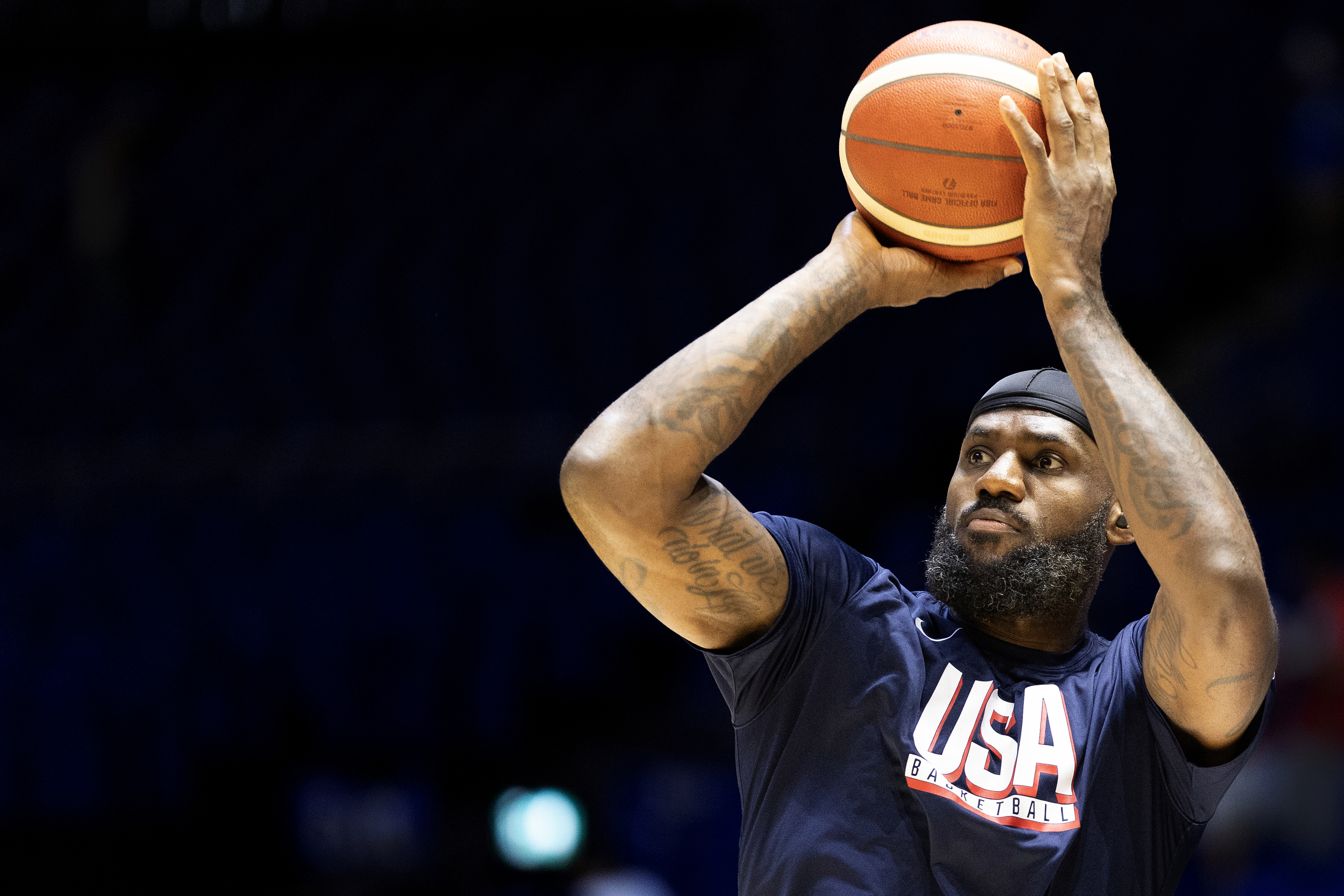 LeBron James, wearing a black headband and USA Basketball shirt, holds a basketball above his head, preparing to take a shot.