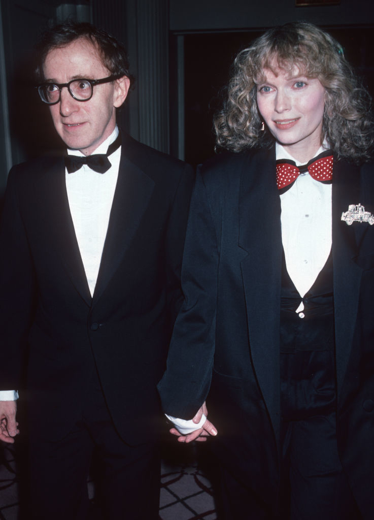 Woody Allen and Mia Farrow on a formal event's red carpet, both wearing tuxedos. They are holding hands