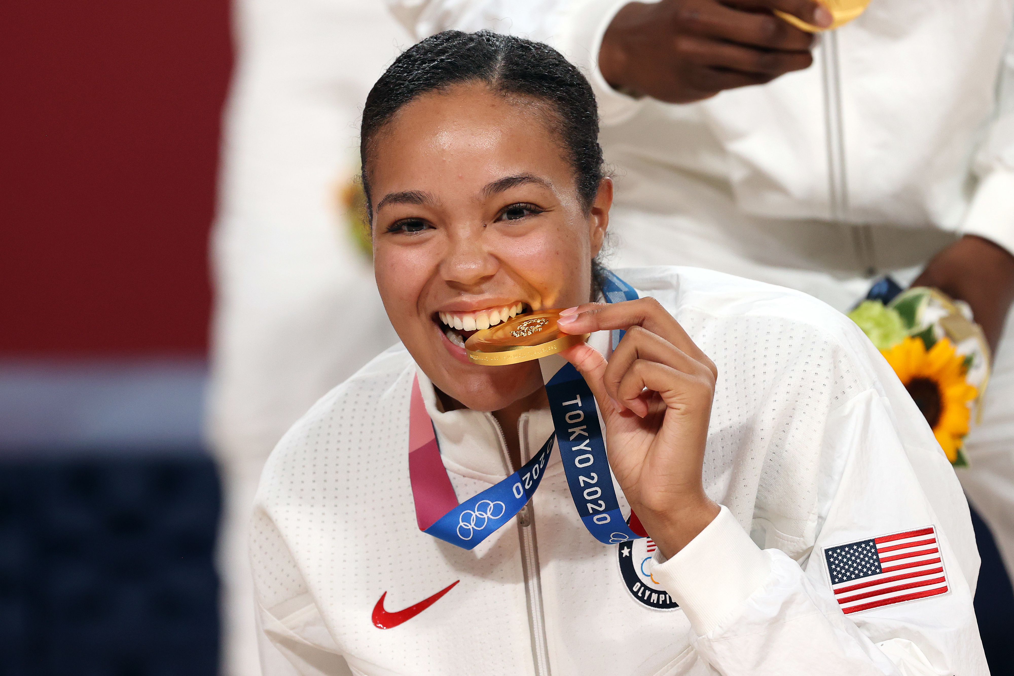 Olympian biting gold medal, wearing white Team USA track suit at Tokyo 2020