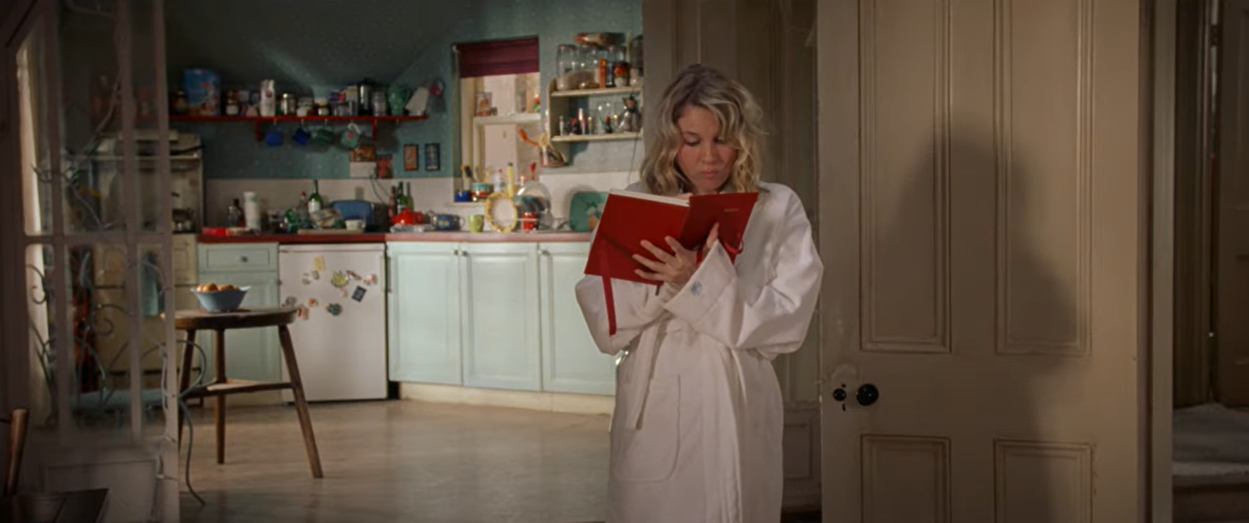 Renée Zellweger reads a red book in a kitchen wearing a white bathrobe, a scene from &quot;Bridget Jones's Diary&quot;